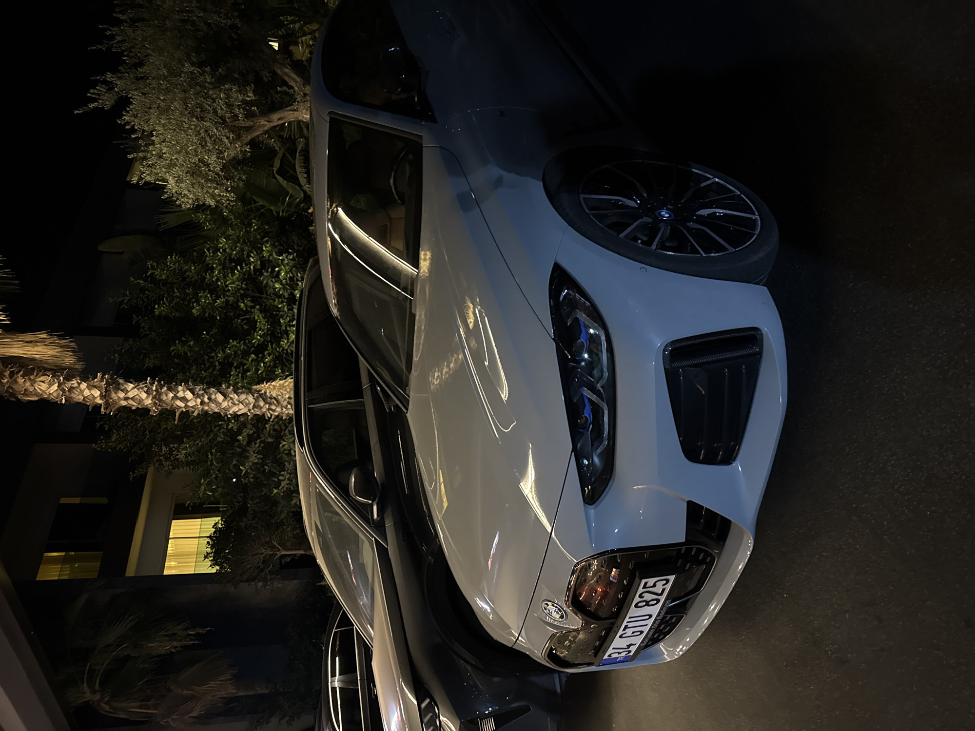 This is a nighttime, eye-level, medium shot of two parked cars, likely in Bodrum, Turkey, given the license plate. In the foreground, a light-colored BMW sedan is parked on asphalt. The car's front is angled towards the viewer, showing its grille, headlights, and license plate which reads "34 GTU 825". Behind it, a darker SUV, possibly also a BMW, is parked. The background reveals a building with illuminated windows and lush green foliage, including palm trees and an olive tree. The lighting is dim, with artificial lights from the building and the cars illuminating the scene. There are no people visible in the image.
