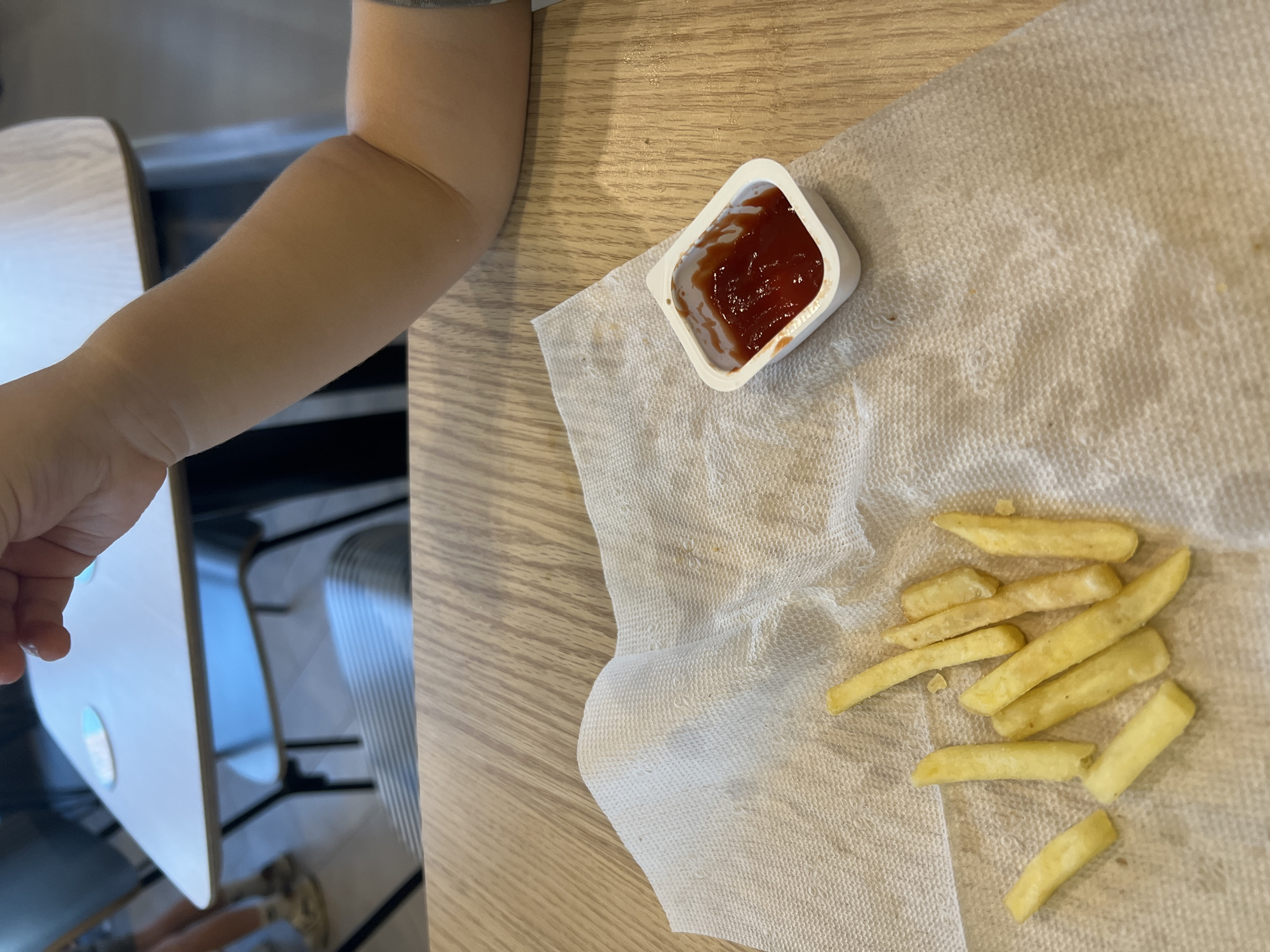 The image shows a close-up view of a child's arm resting on a wooden table, next to a white paper napkin with french fries and a small container of ketchup. The child's arm is light-skinned and appears to be that of a young child. The french fries are golden brown and scattered on the napkin. The ketchup is a deep red color and is contained in a small, white, square plastic cup with a slightly crumpled edge. The table appears to be made of light-colored wood with a visible grain. In the background, out of focus, there are hints of furniture, possibly chairs, and a reflective surface, perhaps a table or a high chair tray. The lighting suggests it is daytime. The scene is casual and appears to be a mealtime setting. There is no visible text in the image.