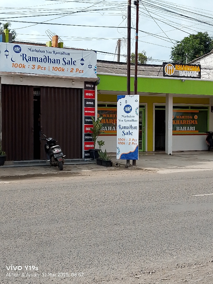The image shows a street scene in Jatibarang, Indonesia. The street is paved with grey asphalt and is bordered by two businesses on either side. Both businesses have signs advertising a "Ramadhan Sale" and both have the words "KHARISMA BAHARI" on their signs. The business on the left has a partially open door revealing a cluttered interior with shelves filled with various products. A black motorcycle is parked in front of this business. The business on the right has a closed door and a window revealing a dark interior. The street is empty of people and vehicles. The businesses are painted in a vibrant green and yellow. Overhead wires run across the street and the sky is overcast. The location, based on the coordinates provided, is Jl. Raya Jatibarang - Pamanukan, Jatibarang, Indramayu Regency, West Java 45261, Indonesia.