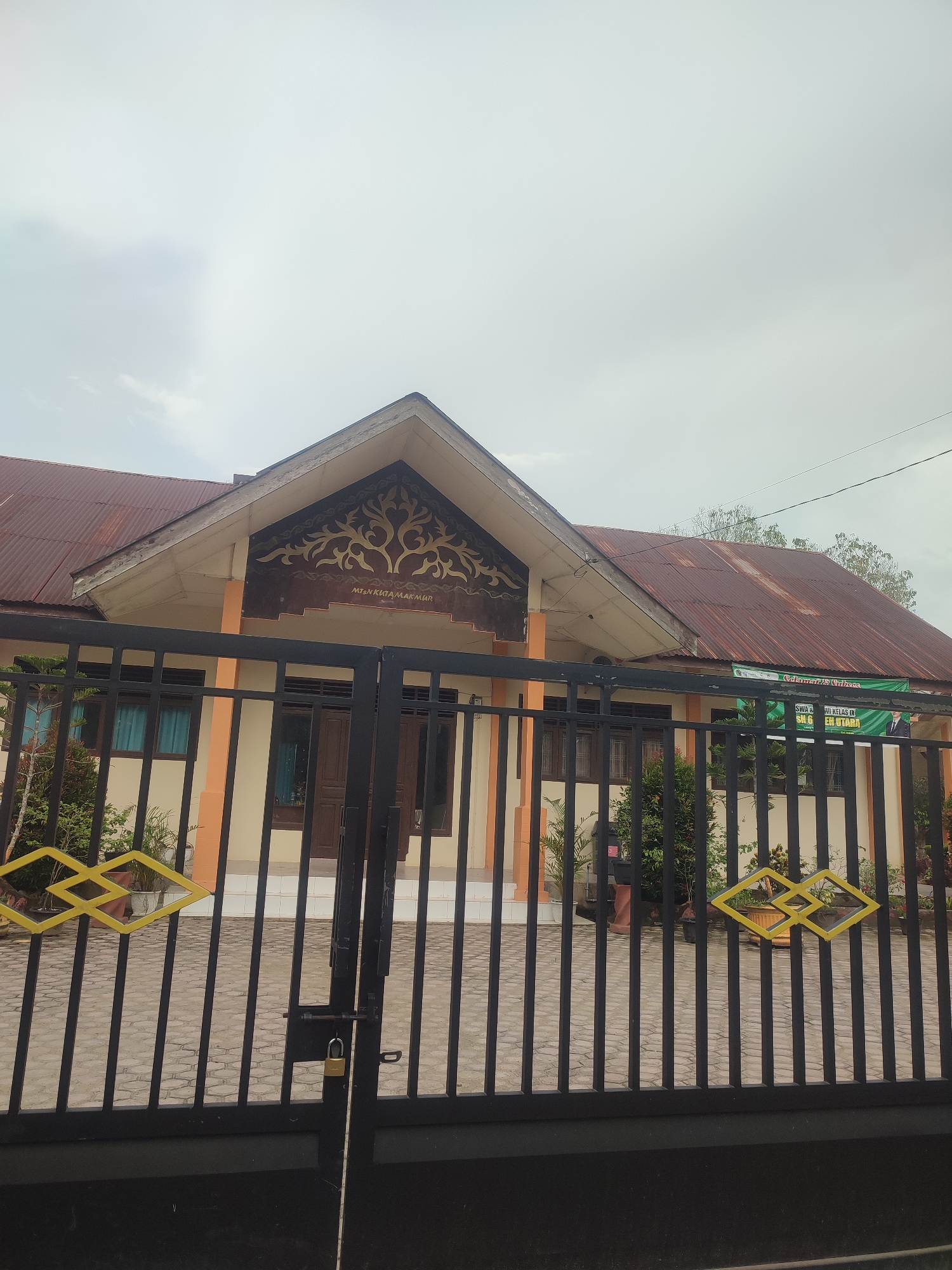 The image shows a single-story building behind a black metal fence. The building is cream-colored with orange accents on the pillars. The roof is red and rusty. Above the entrance is a decorative brown panel with a tree-like design and the inscription "MTsn KUTA MAKMUR". The fence has yellow diamond-shaped decorations. A padlock is visible on the gate. The background consists of a cloudy sky and glimpses of trees. The location is Blangriek, Indonesia, based on the building signage.