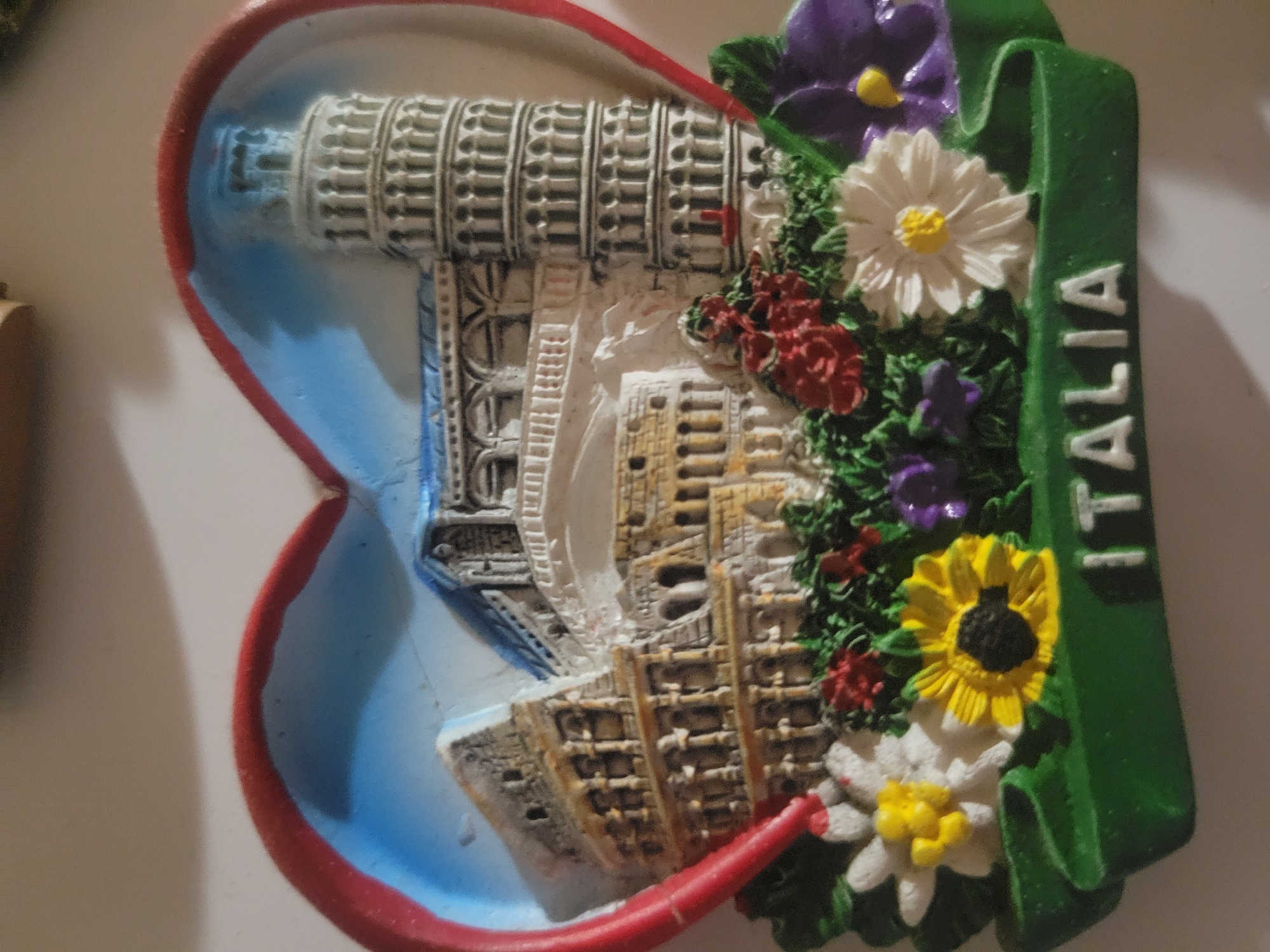 The image is a close-up shot of a souvenir magnet shaped like a heart. Inside the heart, a miniature depiction of Italian landmarks, including the Colosseum and the Leaning Tower of Pisa, is visible. The base of the magnet features a green banner with the word "ITALIA" inscribed in white lettering. The magnet is adorned with artificial flowers in various colors, including yellow, white, red, and purple. The background is a plain, light-colored surface, possibly a table. There are no people or animals present in the image. The scene is static and does not depict any actions or interactions. The image does not provide any information about the location of Belaazyorsk, Belarus.