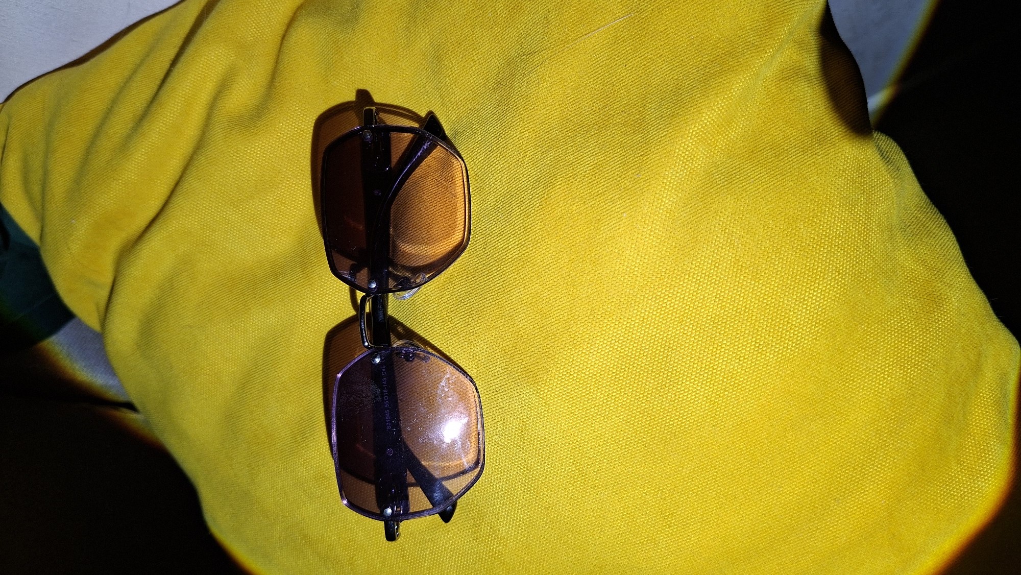 The image shows a pair of sunglasses resting on a bright yellow fabric. The sunglasses have brown lenses and a dark frame. The fabric appears to be textured. The location, Chaikovsky, Russia, is not discernible from the image. There are no people, actions, or interactions visible. The only objects are the sunglasses and the fabric. The environment is dark with a light source illuminating the fabric and sunglasses.