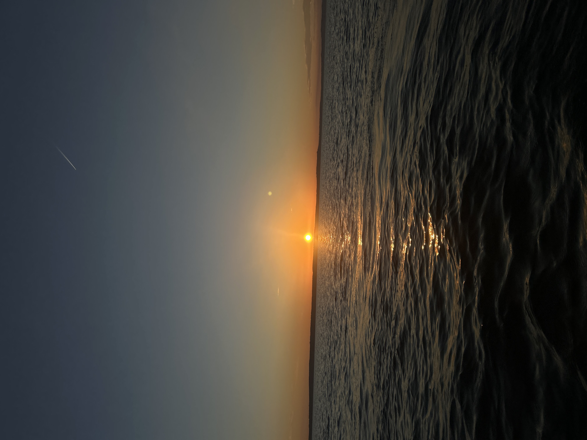 The image depicts a sunset over a body of water, likely a river or lake, with a distant shoreline visible. The sky transitions from a deep blue at the top to a warm orange glow near the horizon where the sun is setting. A thin contrail from an aircraft is visible in the upper right portion of the sky. The water's surface is rippled, reflecting the golden light of the setting sun, creating a shimmering path across the scene. The foreground of the water is darker, with the waves creating shadows and highlights. There are no people or visible man-made structures besides the distant contrail. This scene is consistent with a view of the Kama River near Chaikovsky, Russia, a city known for its hydroelectric power station and its location on the river.