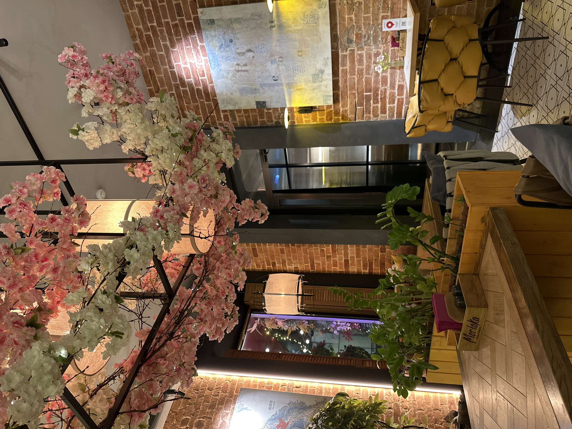The image shows the interior of a restaurant. A large faux cherry blossom tree with pink and white flowers dominates the left side of the frame. The restaurant has brick walls, decorated with artwork. There is an opening or doorway leading to another area. Seating includes a mustard yellow chair with a quilted pattern. There are plants in wooden planters. The planter closest to the camera has the word "Morioka" printed on a block. The location is Ovoshchnoy, Russia.