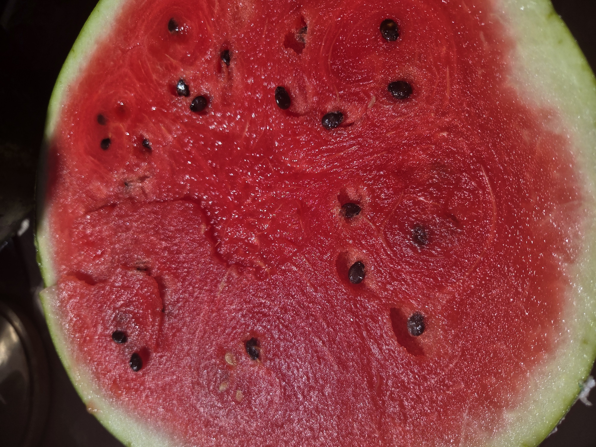 A close-up, overhead shot of a ripe watermelon slice reveals its vibrant red flesh, dotted with black seeds. The juicy texture of the fruit is evident, with glistening droplets of moisture scattered across its surface. The rind, a thin green band, frames the bright red interior. The background is dark and indistinct, suggesting the watermelon is likely indoors, perhaps in a kitchen or on a table. There are no people present, and the scene appears static and undisturbed. The image is likely a food photograph, intended to showcase the freshness and appeal of the watermelon. The location, Восход Снт (фролово), Russia, is provided, but the image itself does not contain any visual cues to corroborate this information.