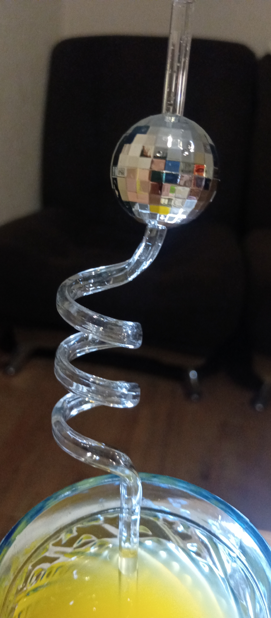 The image shows a clear, spiral drinking straw with a small disco ball attached to the top. The straw is inserted into a glass filled with a yellow liquid, likely a beverage. The background is out of focus, showing a dark colored chair and the top portion of a wood colored floor.