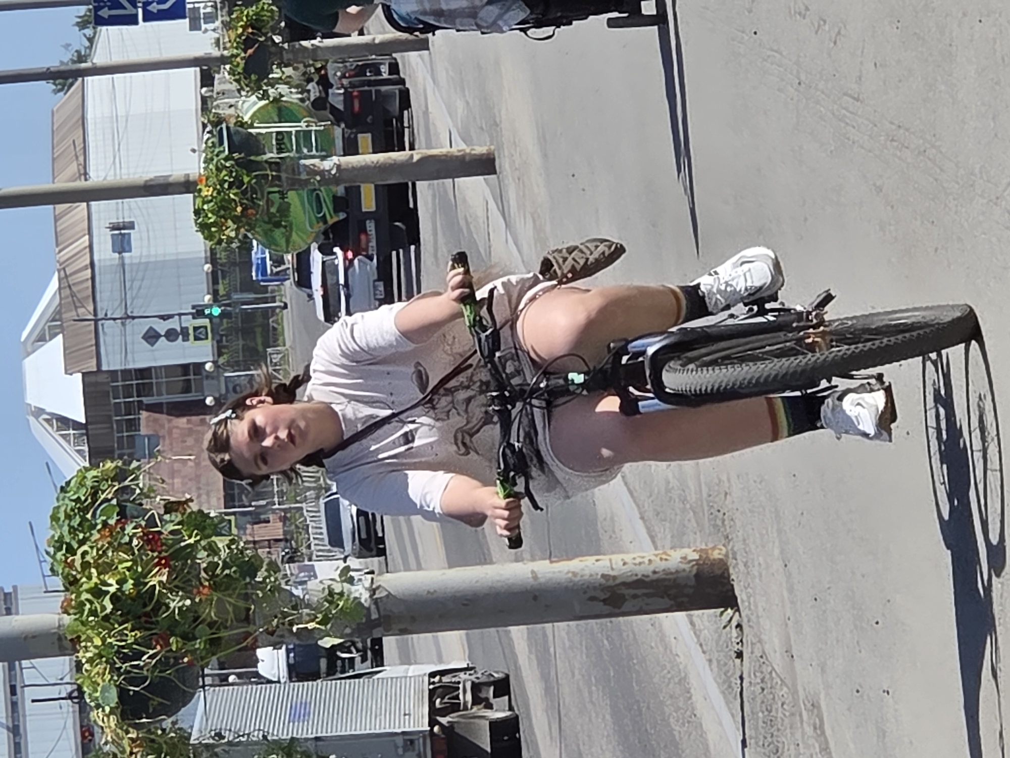 A young woman with brown hair pulled into a ponytail and wearing a white t-shirt and light-colored shorts is riding a bicycle on a paved path. She is looking directly at the camera. She is wearing white sneakers with rainbow-striped socks and has a small brown patterned bag clipped to her shorts. The bicycle has green handlebars and a black frame. In the background, there are vehicles, including trucks and cars, on a road, and buildings. There are also trees with hanging flower baskets on poles along the path. The scene is set in an urban environment on a sunny day.