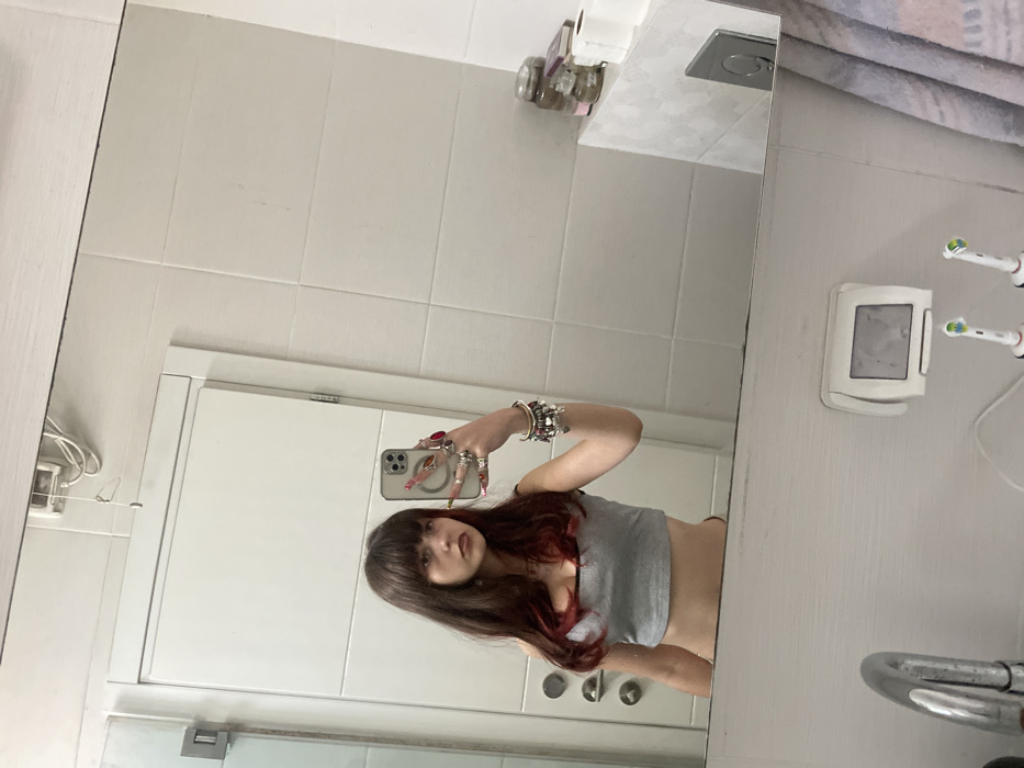 A selfie in a bathroom mirror shows a woman with long brown hair, bangs, and red highlights. She wears a gray tank top and numerous rings and bracelets. The bathroom has white tiled walls, a toilet paper holder, and electric toothbrushes. The reflection captures a white door with a handle and a partial view of a sink area. The mirror is rectangular and positioned above the sink.