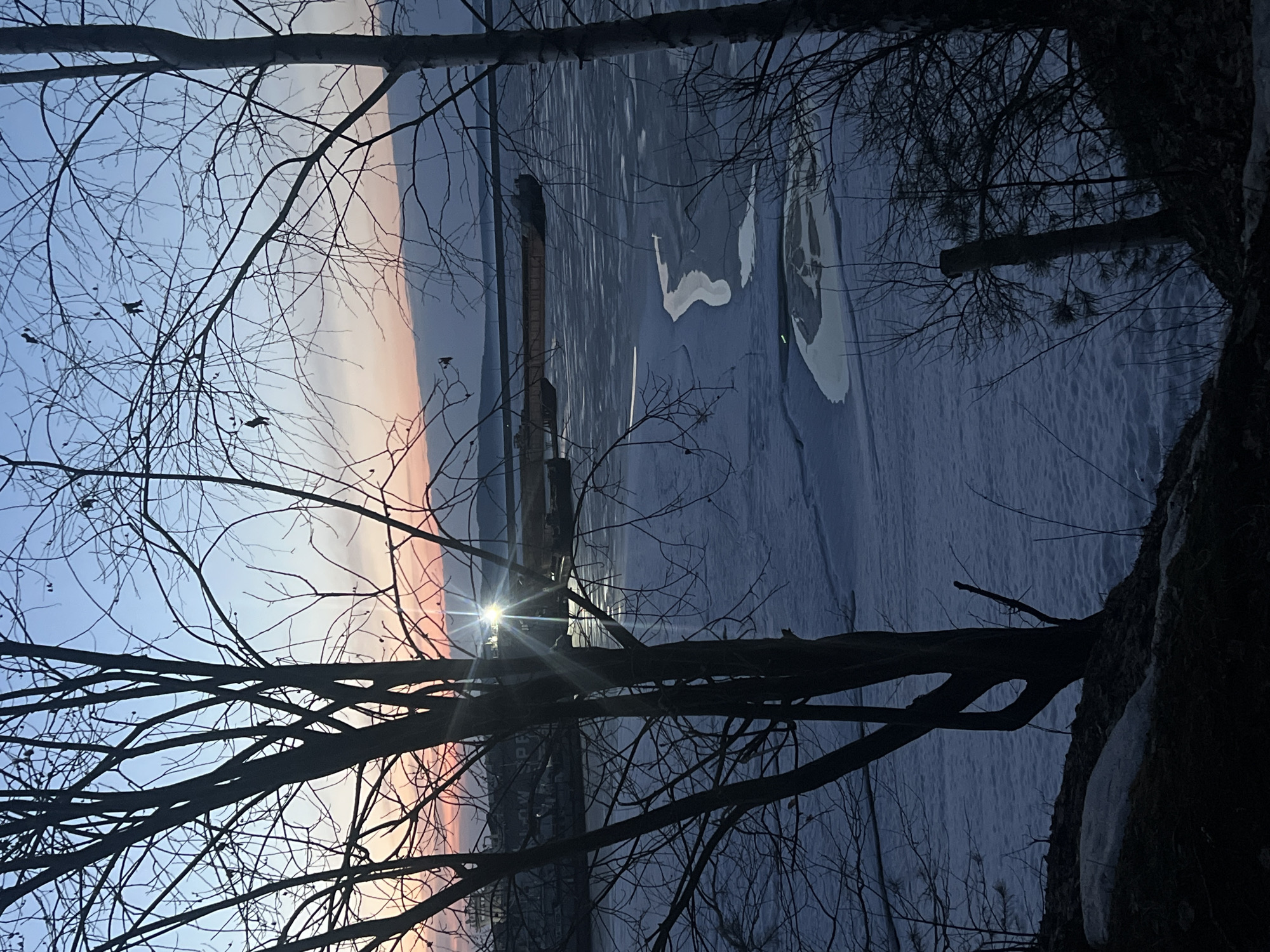 The image shows a winter scene near the Kama River in Chaikovsky, Russia. The foreground features bare tree branches silhouetted against a sky transitioning from blue to pink. The river, partially frozen, occupies the middle ground, with some areas of open water or thin ice visible. An industrial structure, potentially a factory chimney, is on the river's edge. The sun appears to be setting, creating a lens flare effect. The scene is captured at an angle, suggesting the photographer may be situated on a hillside or elevated position.