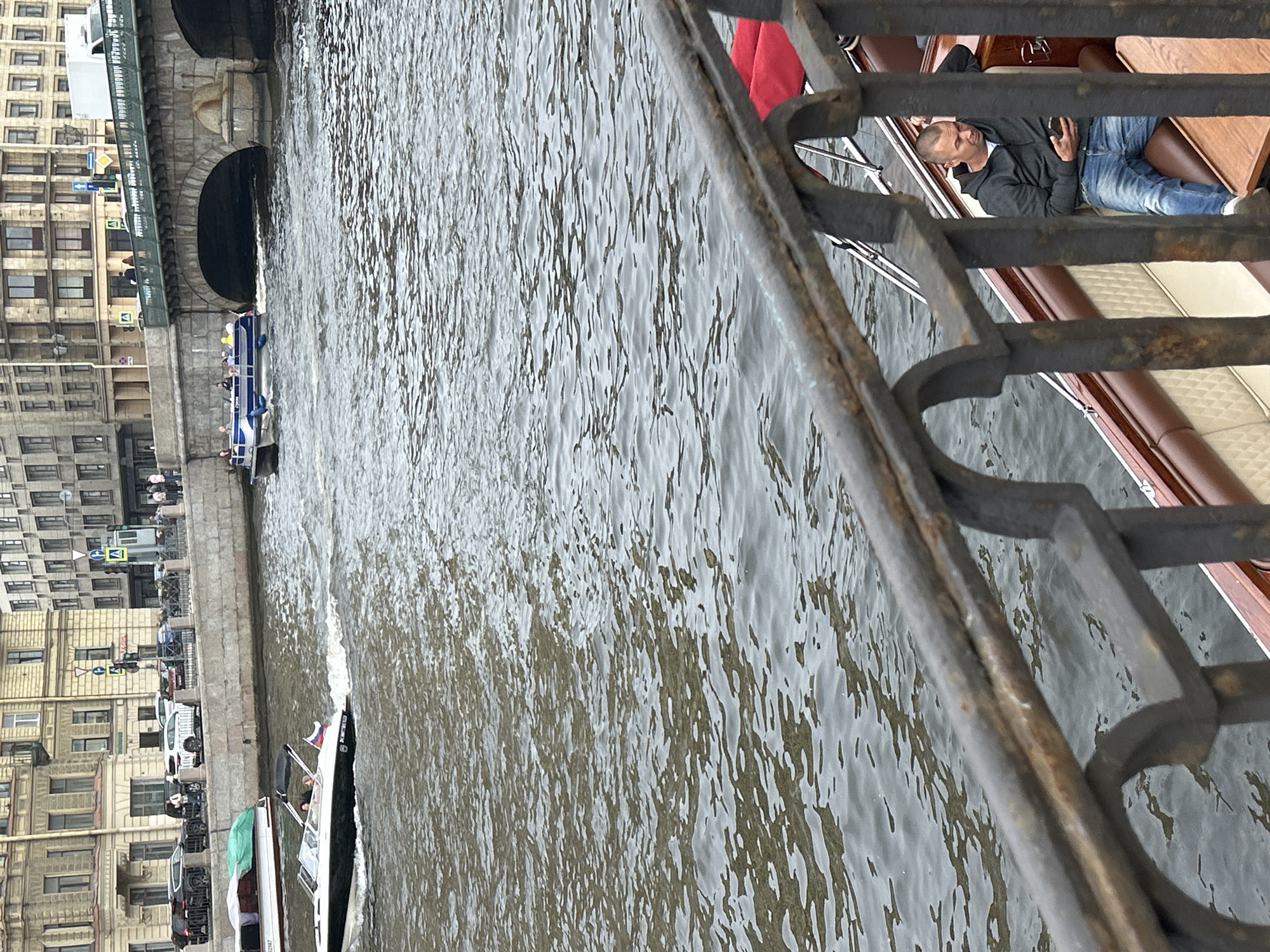 The image shows a body of water, likely a river or canal, with several boats on it. In the foreground, there is a dark metal railing that partially obscures the view. On the water, there are a few boats. One boat in the middle ground is white with blue accents and appears to be a tour boat, with several people visible inside. Another boat, a smaller white one with a green canopy, is docked in the foreground. In the background, a larger boat with a red tarp is visible, with a man in a dark hoodie and jeans lying down on it.
To the left of the water, there is a cityscape with buildings lining the waterfront. The architecture suggests an older European city, with ornate details on the buildings and arched bridges. The buildings are tall and multi-story, with many windows. There are also signs visible on the buildings and street level, including what appears to be a directional sign with a blue square and white arrow, and another sign with a blue letter "M" inside a yellow square. Cars are parked along the waterfront.
The water is choppy, indicating some movement, and appears to be a dark, murky color. The sky is overcast, suggesting a cloudy or rainy day. The time of day is not definitively ascertainable, but the lighting conditions suggest it could be daytime. The overall mood is somewhat subdued, perhaps due to the weather. One boat has a Russian flag, providing a clue to the location. There is some text visible on the back of the small white boat, but it is blurry and unreadable.