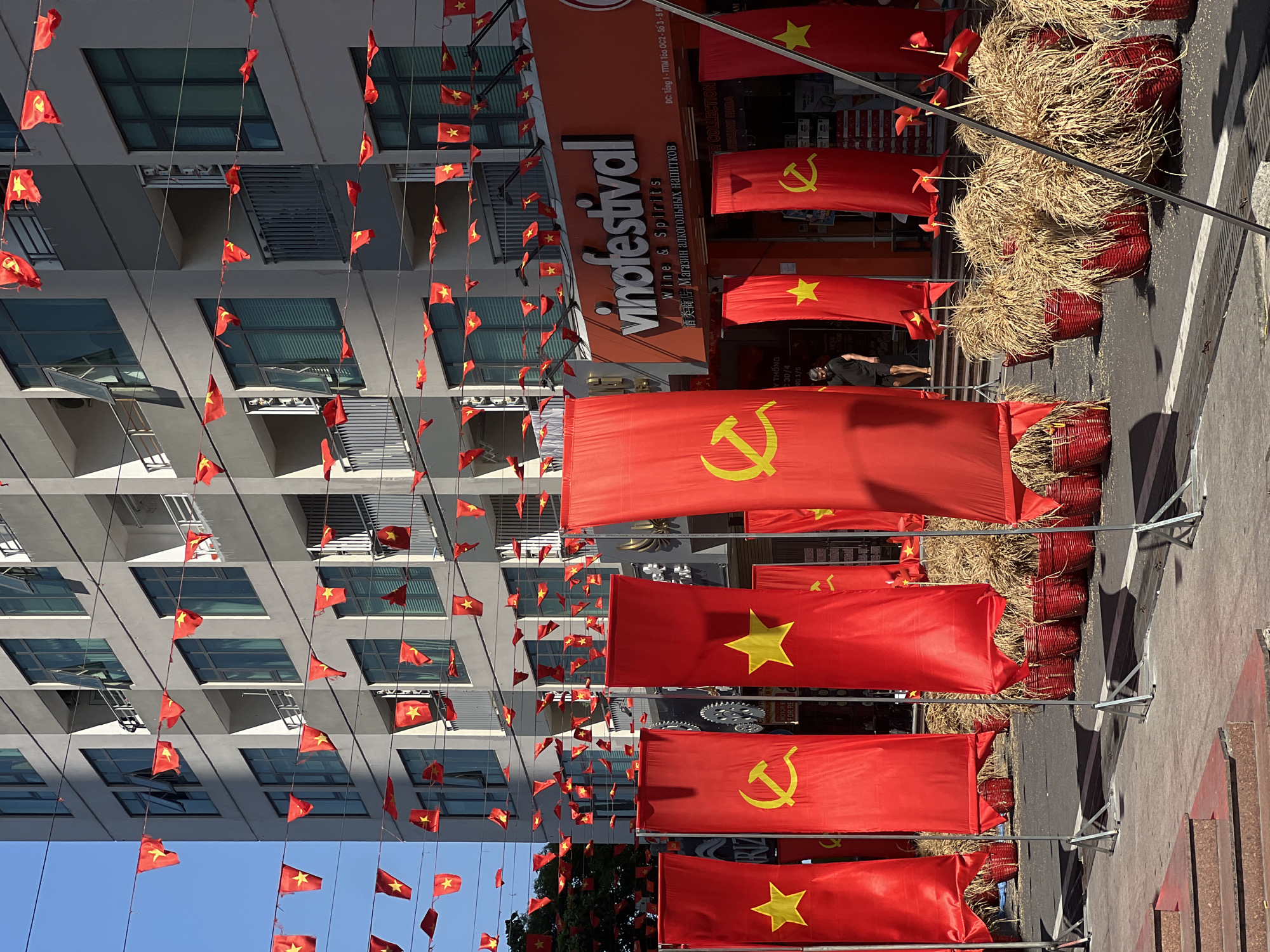 A vibrant street scene in Bắc Nha Trang, Vietnam, is captured during a sunny daytime. The street is heavily decorated with numerous red flags, creating a festive atmosphere. Large vertical banners line one side of the street; some display the yellow five-pointed star of the Vietnamese national flag, while others feature the yellow hammer and sickle symbol of the Communist Party of Vietnam. Below these banners, red woven baskets filled with straw are placed as decorations.
In the middle ground, a large orange sign advertises "vinofestival Wine & Spirits," with additional text in Chinese ("酒类商店") and Russian ("Магазин алкогольных напитков"), both translating to "alcohol shop." The sign also includes Vietnamese address details: "ĐC: Tổng 1 - TTTM Tòa OC2 - Số 3-5 P." Another visible text fragment includes "30/4" and "1/5," likely referring to April 30th (Reunification Day) and May 1st (International Workers' Day), suggesting the decorations are for these national holidays.
In the background, a modern multi-story building with many windows and balconies is visible. Strings of smaller Vietnamese national flags are strung across the street, adding to the celebratory decor. A person wearing a black shirt and face mask is seen near the flag displays. The clear blue sky and bright sunlight indicate good weather conditions. The scene depicts a lively street preparing for or celebrating an event, possibly a wine festival, amidst national festivities.