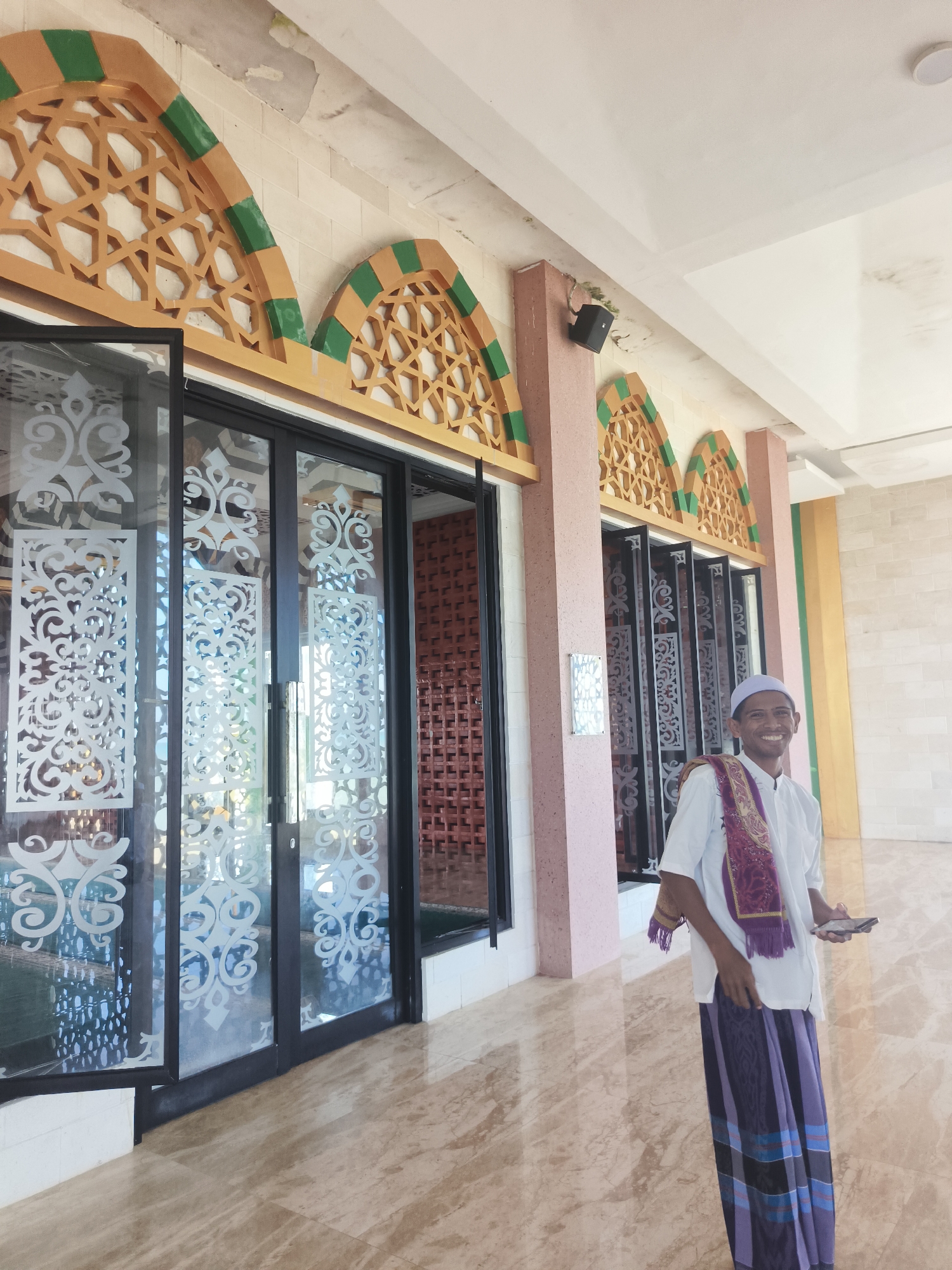 A young man is standing on a marble floor in front of a building with a pink facade and intricately designed arched windows. He is wearing a white shirt, a white cap, a purple and gold patterned sash, and a purple sarong. He is smiling and holding a mobile phone in his right hand. The building appears to be a mosque, with its decorative elements and architectural style suggestive of religious architecture in Indonesia. The windows are large and made of glass with decorative white stencils. The floor is made of polished marble with a marbled pattern. The lighting suggests it is daytime, with sunlight streaming in from the left. The background shows more of the building's facade, including more arched windows and a red brick wall visible through the glass. The man is the sole human subject in the frame. The overall scene is clean and well-maintained.