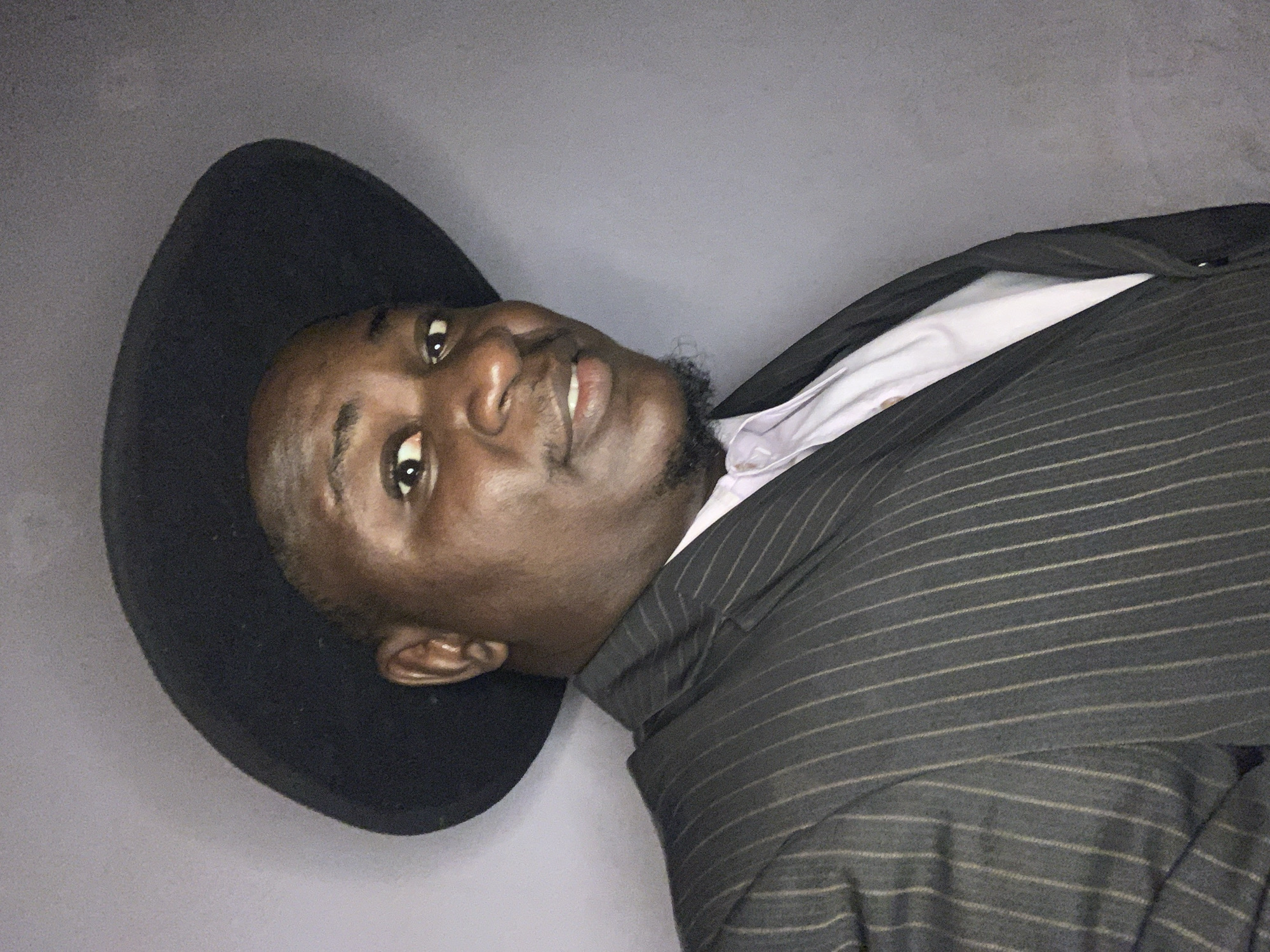 This image depicts a dark-skinned male subject in a close-up portrait, reportedly taken in Akwanga, Nigeria. The subject, positioned centrally, wears a black, wide-brimmed fedora hat that partially covers his forehead. He has dark eyes that meet the camera's gaze, a prominent nose, and a faint smile with closed lips. A short, dark beard and mustache frame his mouth. He is dressed in a dark gray pinstripe suit jacket featuring thin, light vertical stripes, worn over a light-colored, possibly pale purple or white, collared shirt. The left shoulder and lapel of the suit are visible, indicating the jacket is buttoned or partially open. The background is a uniform, plain gray wall, devoid of any discernible features or textures. The lighting is consistent, casting no harsh shadows, and illuminates the subject's face and upper torso directly.