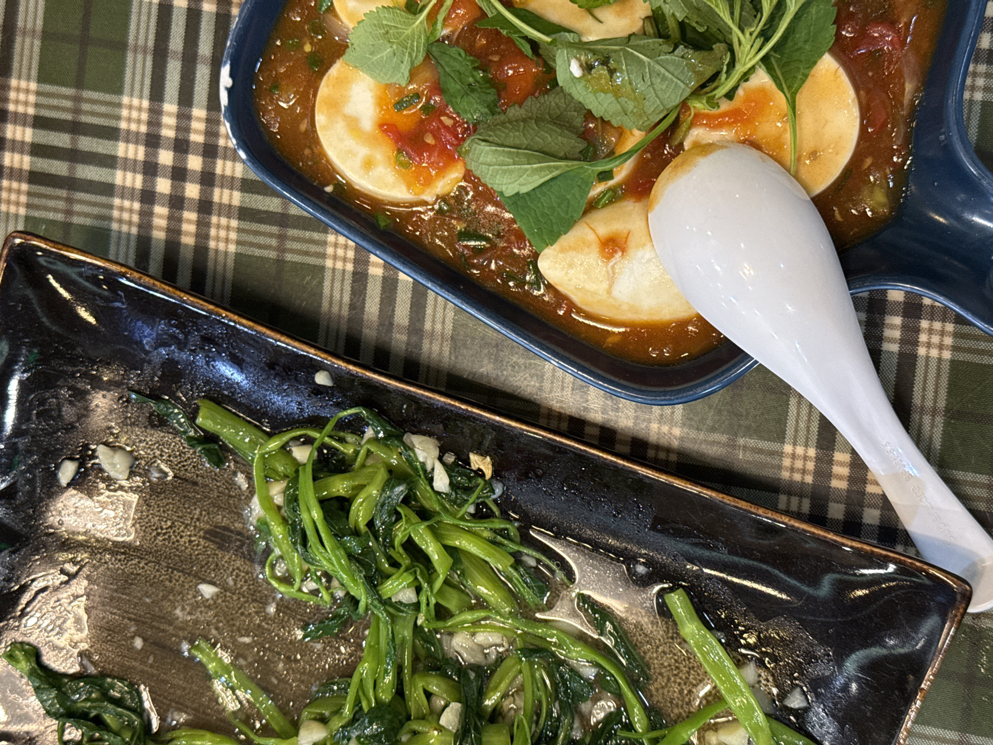 The photograph, taken from a top-down perspective, features two dishes on a tablecloth with a green and brown checkered pattern.
In the upper right quadrant, a blue rectangular dish contains circular pieces of what appears to be tofu or eggs, arranged in a brown sauce with visible chunks of tomato. Fresh green herbs, resembling mint or basil, are scattered on top. A white ceramic spoon rests in the dish, angled diagonally towards the lower right.
Below, a black rectangular plate holds a pile of bright green vegetables that appear to be stir-fried with garlic. The plate has a glossy finish that reflects light, and a thin brown rim around its edge. The tablecloth is visibly stained around the food dishes.
Given the presence of what appears to be tofu with a tomato sauce and stir-fried morning glory, the setting may be a restaurant in Hanoi, Vietnam.