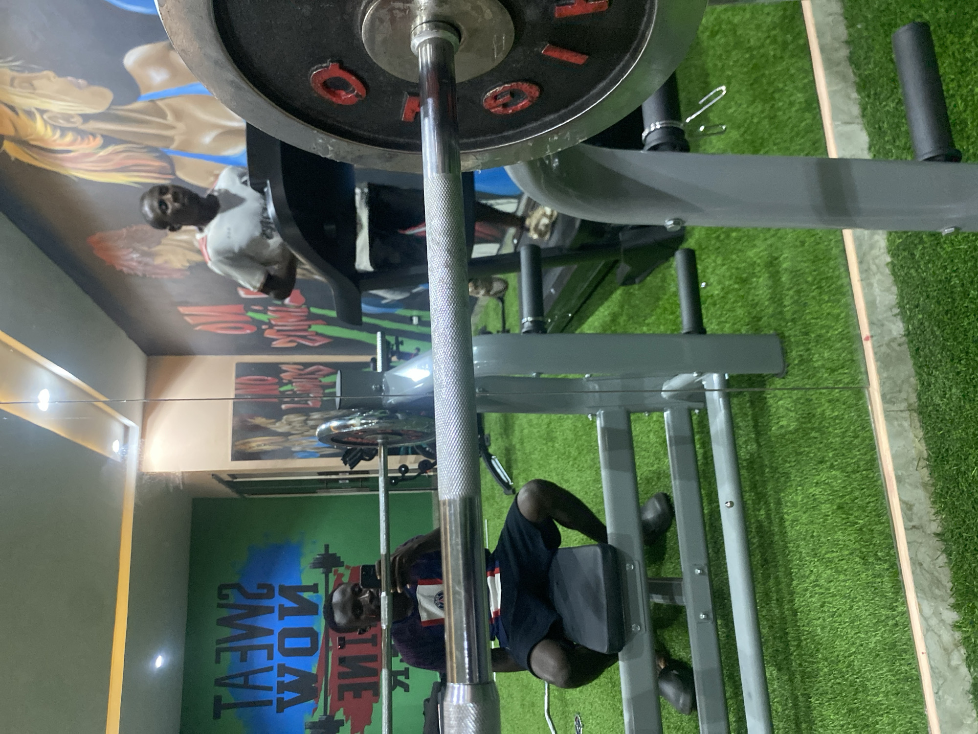 In Barden Yana, Nigeria, a man is seen in a gym. He is seated on a weight bench, holding a smartphone to take a selfie. Behind him, another man is on a treadmill, running. The gym features green turf flooring and a wall mural with motivational text and artwork of a woman lifting weights. A barbell with weights is prominently positioned in the foreground, reflecting in the mirror.