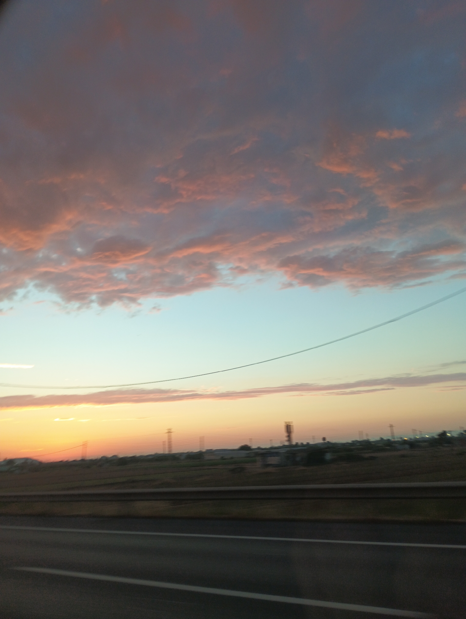 A horizontal, low-angle, medium shot captures a wide expanse of sky and a hint of the ground from the perspective of a moving vehicle. The sky dominates the frame, transitioning from a dusky, pinkish-orange hue near the horizon to a soft, pale blue overhead, eventually darkening to a deep gray with scattered pink clouds in the upper portion. A thin, dark power line stretches diagonally across the mid-section of the sky. Below the sky, a strip of land is visible, appearing as a blurred, brown expanse with sparse vegetation and industrial structures or utility poles scattered across it. In the foreground, the asphalt road with white lane markings is visible, suggesting the image was taken from inside a car. The overall impression is of a rural or semi-industrial landscape at dusk or dawn, viewed from a roadway. The location is described as Unknown, Unknown.