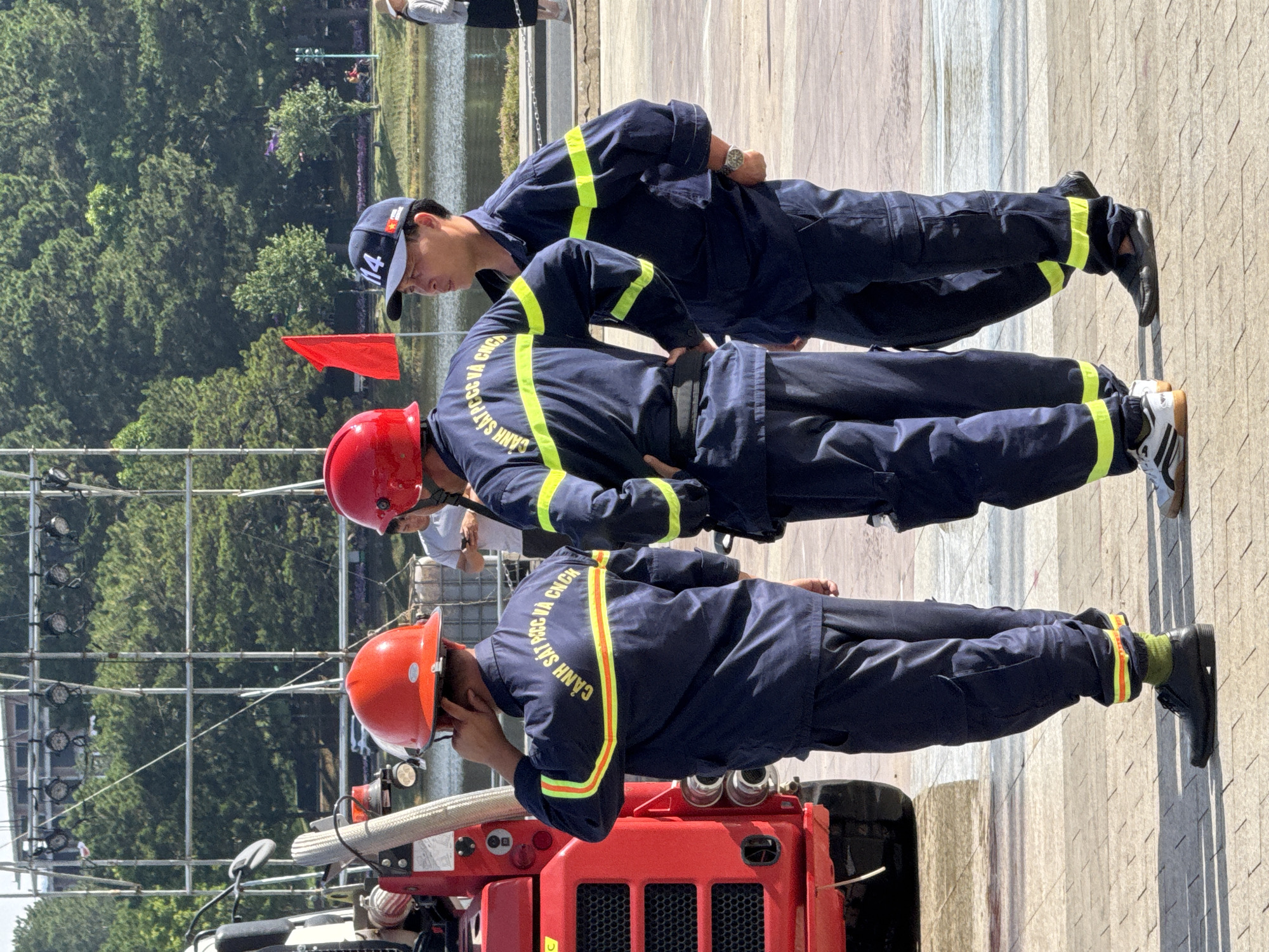 The image depicts three individuals in dark blue uniforms with yellow and orange reflective stripes, likely firefighters, standing outdoors. The person on the left wears a red helmet and has their hand to their ear. The central figure also wears a red helmet. The person on the right is wearing a dark blue cap with the number "14" prominently displayed and is looking to the right. They are positioned near a large red vehicle with a hose attached, which is partially visible. In the background, there are trees, a body of water, and a distant structure. A red flag is visible behind the central figure. The ground appears to be paved with square tiles. The scene is set in an outdoor environment that resembles a public area, possibly a park or event space. The presence of trees and a lake suggests a location like Xuan Huong Lake in Da Lat, Vietnam, known for its scenic beauty and occasional public events.