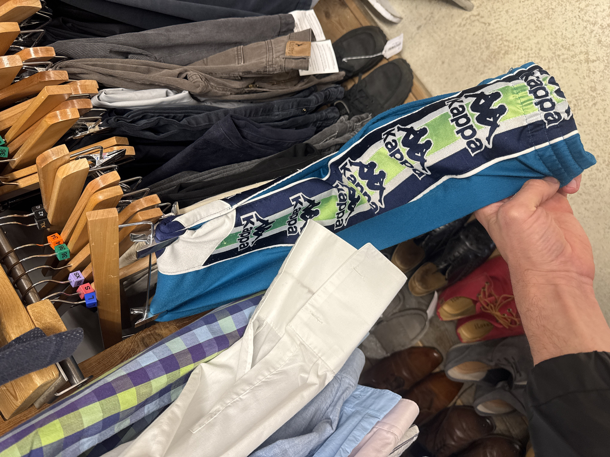 A person's right hand holds up a pair of blue tracksuit bottoms featuring prominent "Kappa" branding with white, green, and black stripes running down the side. The "Kappa" logo, depicting two silhouettes, and the brand name are repeated along this stripe.
The scene appears to be inside a clothing store, possibly a thrift shop or a large retail outlet, given the diverse collection of garments and footwear. In the background and mid-ground, numerous clothes are visible, stacked or hanging on wooden hangers with metal clips. These include various pairs of trousers or pants in shades of grey, brown, and black, as well as folded shirts in white, plaid, light blue, and pink. One brown garment label is partially visible, showing the text "LLOYD'S".
Several plastic size tags are attached to the hangers, displaying "XL", "42", "36", "S", "XS", and "L". Other small tags or labels show numbers like "36", "3614", and "34". On the floor below the clothing racks, an assortment of shoes is scattered, including red boots, brown dress shoes, and grey and tan casual footwear. The overall lighting is artificial and suggests an indoor setting. The person's hand and lower forearm are visible, actively engaging with the clothing.