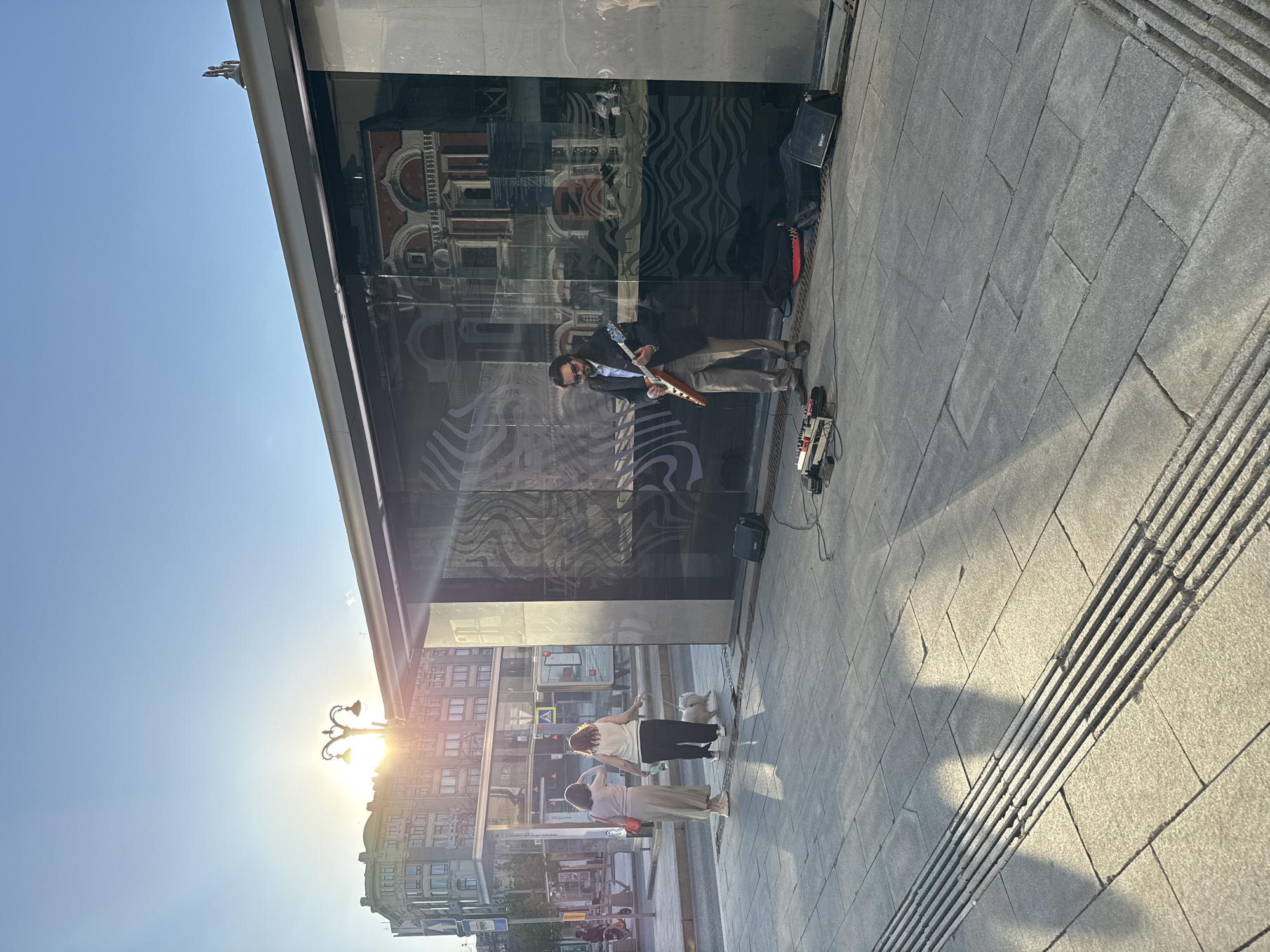 A man with long hair and dark glasses plays an electric guitar on a sidewalk near a building with large, reflective windows. A black amplifier and effects pedals are next to him. Two women and a small white dog stand on the sidewalk near the building. The sun shines brightly in the background, illuminating the buildings across the street. The location is near Manezhnaya Square in Moscow, Russia, as reflected in the building's windows.