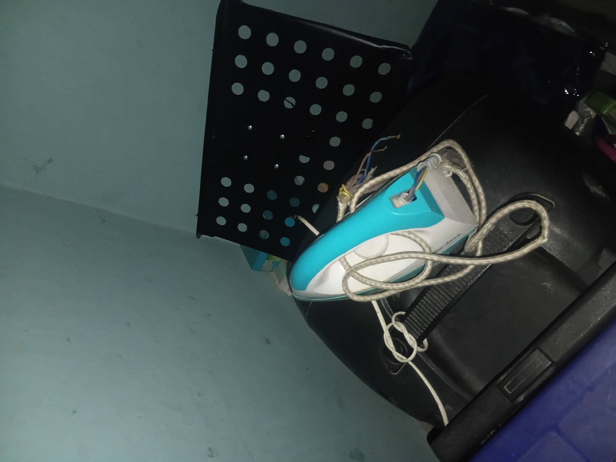 A turquoise and white iron sits on a black, possibly leather, surface. The iron's power cord is coiled around it. Behind the iron is a black object with a grid of circular holes. The background is a pale blue wall. The scene is dimly lit, suggesting it is indoors. The overall impression is of a storage area or a corner of a room, possibly in Jalingo, Nigeria.
