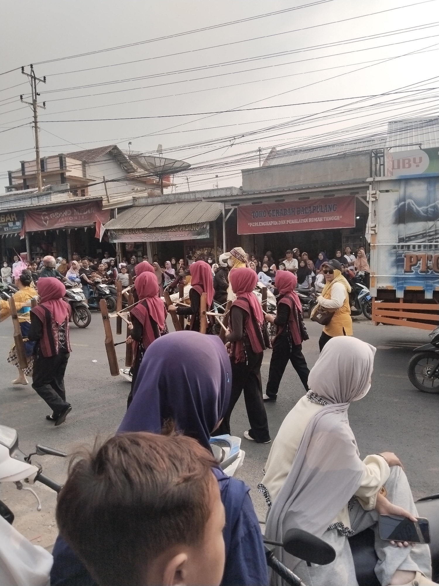In Balapulang Kulon, Indonesia, a street parade is underway, captured from a low angle. Several young women, dressed in traditional attire with maroon headscarves and patterned tunics, are marching and carrying bamboo instruments. They are accompanied by spectators lining the street, some on motorcycles. The background features buildings, banners for local businesses like "GRAND MEDIA STATIONERY" and "TOKO GERABAH BALAPULANG," and overhead power lines, indicating an urban or semi-urban setting. The atmosphere appears to be festive and communal, with participants and observers engaged in the event.