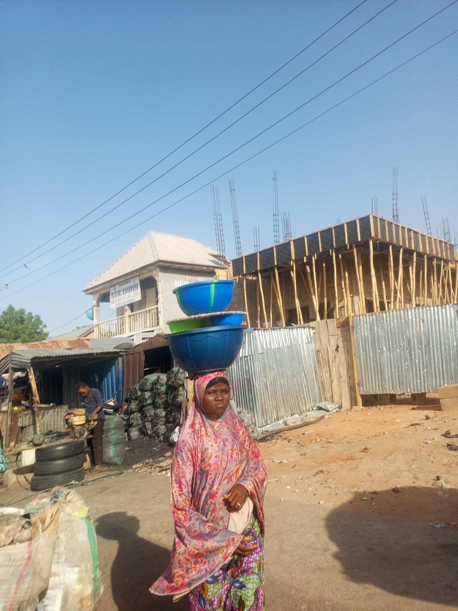 A woman carries three plastic bowls on her head in Tempure, Nigeria. The bowls are blue, green, and blue. She wears a pink, red, and green patterned shawl and a multicolored patterned skirt. In the background are two buildings: one is a two-story building with a sign that reads "MEDICAL DIAGNOSTIC." The other building is under construction with wooden beams and metal rebar extending from the top. A corrugated metal fence stands in front of the construction site. To the left, a man stands near a tire shop. The ground is sandy. Power lines stretch across the sky. The sky is blue.