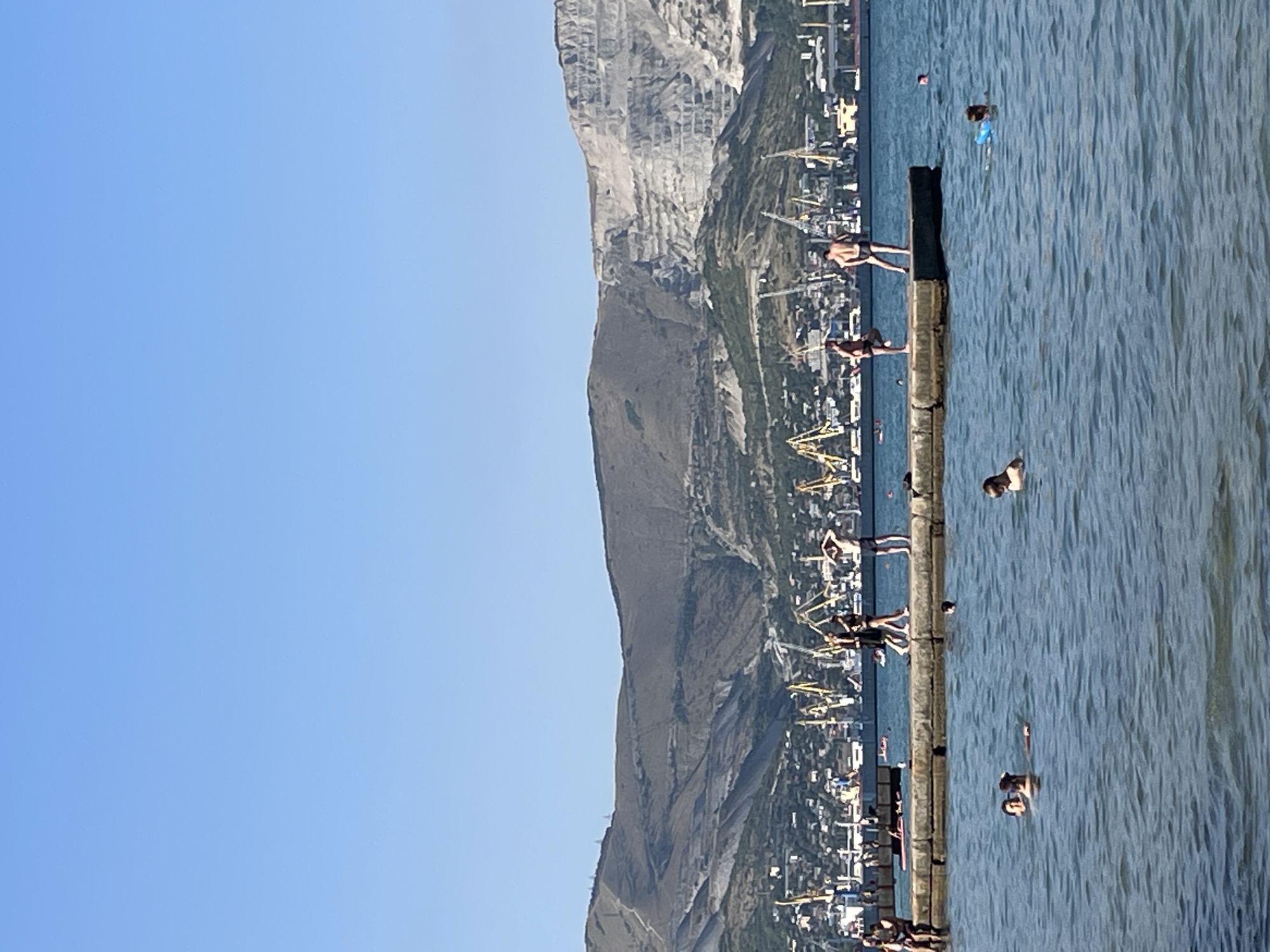 The image depicts a sunny day at a coastal area, likely in Ol'ginskii, Russia, given the context. The scene is dominated by a vast expanse of blue water on the right and a clear blue sky on the left. In the background, a steep, rocky mountain looms over a coastal town with buildings and numerous yellow cranes, suggesting a port or industrial area.
Along a concrete pier extending into the water, several people are visible. Some are standing on the pier, appearing to be enjoying the view or preparing to enter the water. Others are in the water, swimming or wading. The water is calm with gentle ripples. The overall mood appears relaxed and recreational, typical of a summer day at the beach. The time of day is clearly daytime, indicated by the bright sunlight casting shadows. No visible text is present in the image.