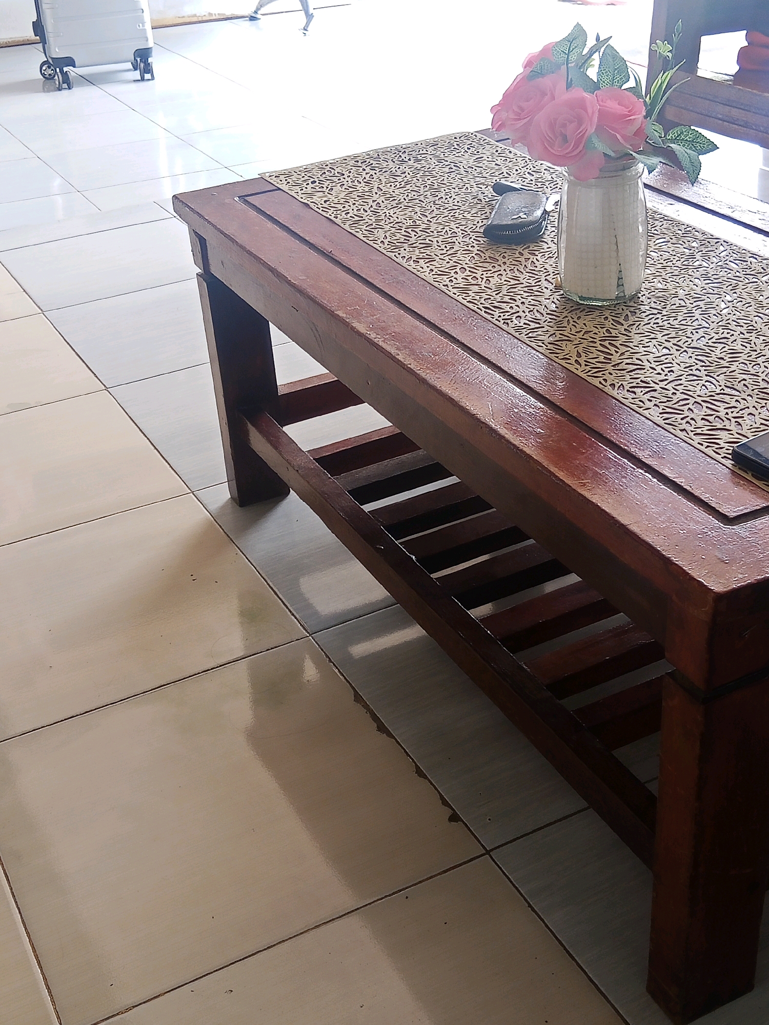 A wooden coffee table with a slotted shelf sits on a tiled floor. A patterned placemat is on top of the table, with a glass vase containing pink roses and a small black pouch next to it. A luggage is in the background. The scene is illuminated by natural light. The image is taken in Lubuklinggau, Indonesia.