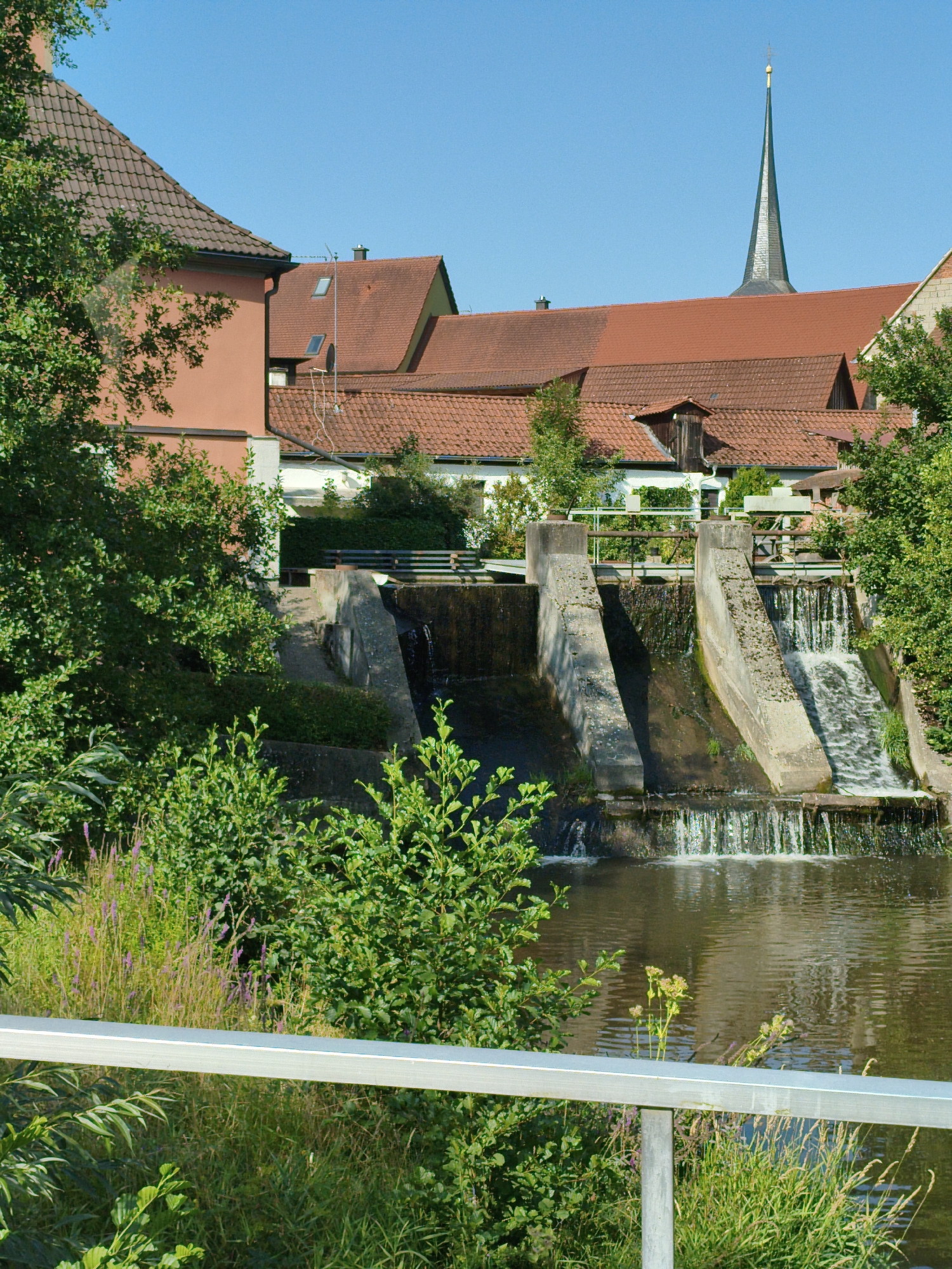 The image depicts a view of the village of Burgwindheim, Germany, focusing on a small dam and the surrounding architecture. A concrete dam structure with water cascading over it dominates the central foreground, with lush green vegetation and trees flanking the water. Behind the dam, buildings with red tile roofs are visible, including what appears to be a church with a tall, slender spire piercing the clear blue sky. The architecture suggests a historical German village setting. The scene is serene, with no people present, and the overall impression is one of a quiet, picturesque rural or small-town environment.