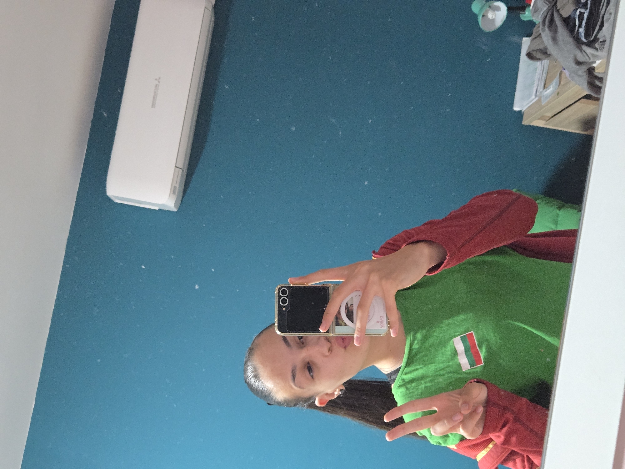 A person is taking a mirror selfie in a room with teal walls. The person is wearing a green shirt with a red and white striped patch, possibly a Bulgarian flag, and a red long-sleeve shirt underneath. They are holding a phone with two visible camera lenses. A white air conditioner unit is mounted high on the wall, while items are visible on a shelf in the upper right corner of the mirror. The location is presumed to be in Pleven, Bulgaria based on the potential flag patch, but the scene lacks specific details to confirm the location definitively.