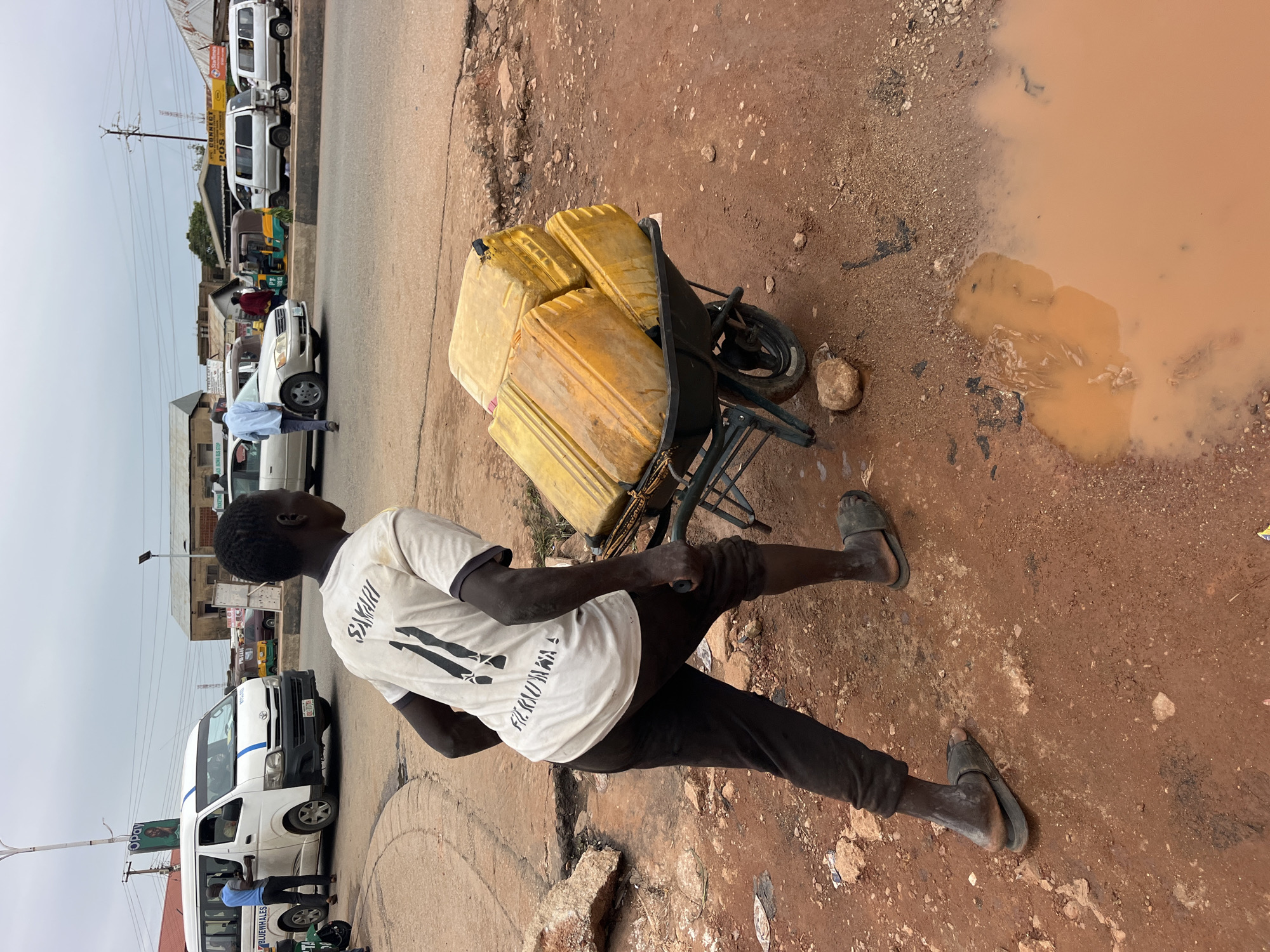 In the city of Unknown, Unknown, on a cloudy day, a young Black male is pushing a wheelbarrow filled with several large yellow jerrycans. He is wearing a white t-shirt with black lettering and graphics, black pants, and dark-colored sandals. The wheelbarrow has a black frame and one wheel. The ground is unpaved, consisting of reddish-brown dirt and gravel, with a large puddle of muddy water in the foreground. In the background, a busy street is visible with several vehicles, including a white minivan with blue stripes, a silver sedan, two white vans, and a small three-wheeled vehicle. Buildings and power lines are also present in the background. The scene appears to be a roadside in an urban or semi-urban environment where water transportation is being done by manual labor.