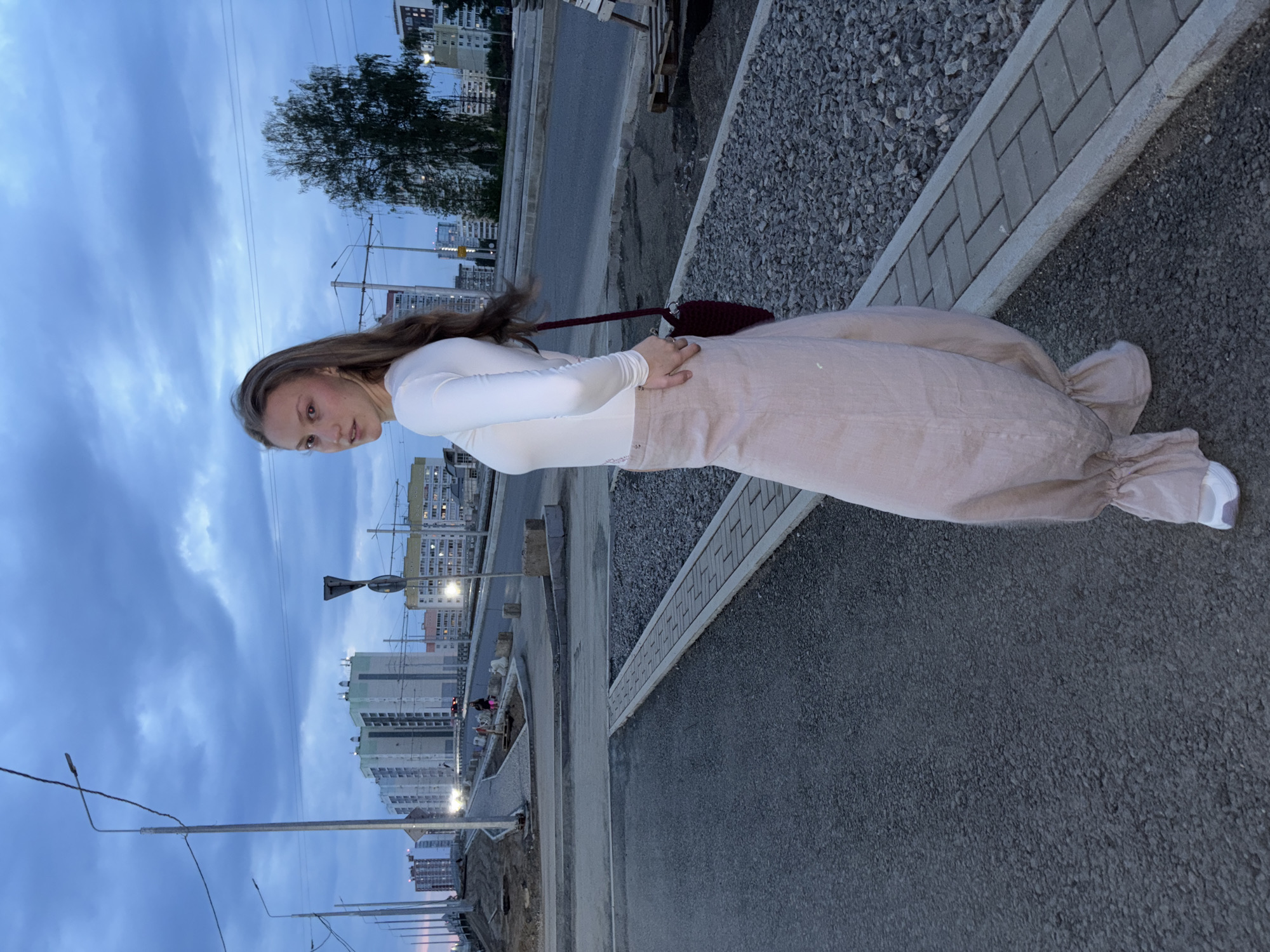 A young woman stands on a paved city street in Perm, Russia, with buildings visible in the background under a cloudy sky. The woman is wearing a white long-sleeved shirt and a light pink skirt. She is carrying a dark burgundy purse. The street is bordered by gravel and concrete dividers. Streetlights are also visible.