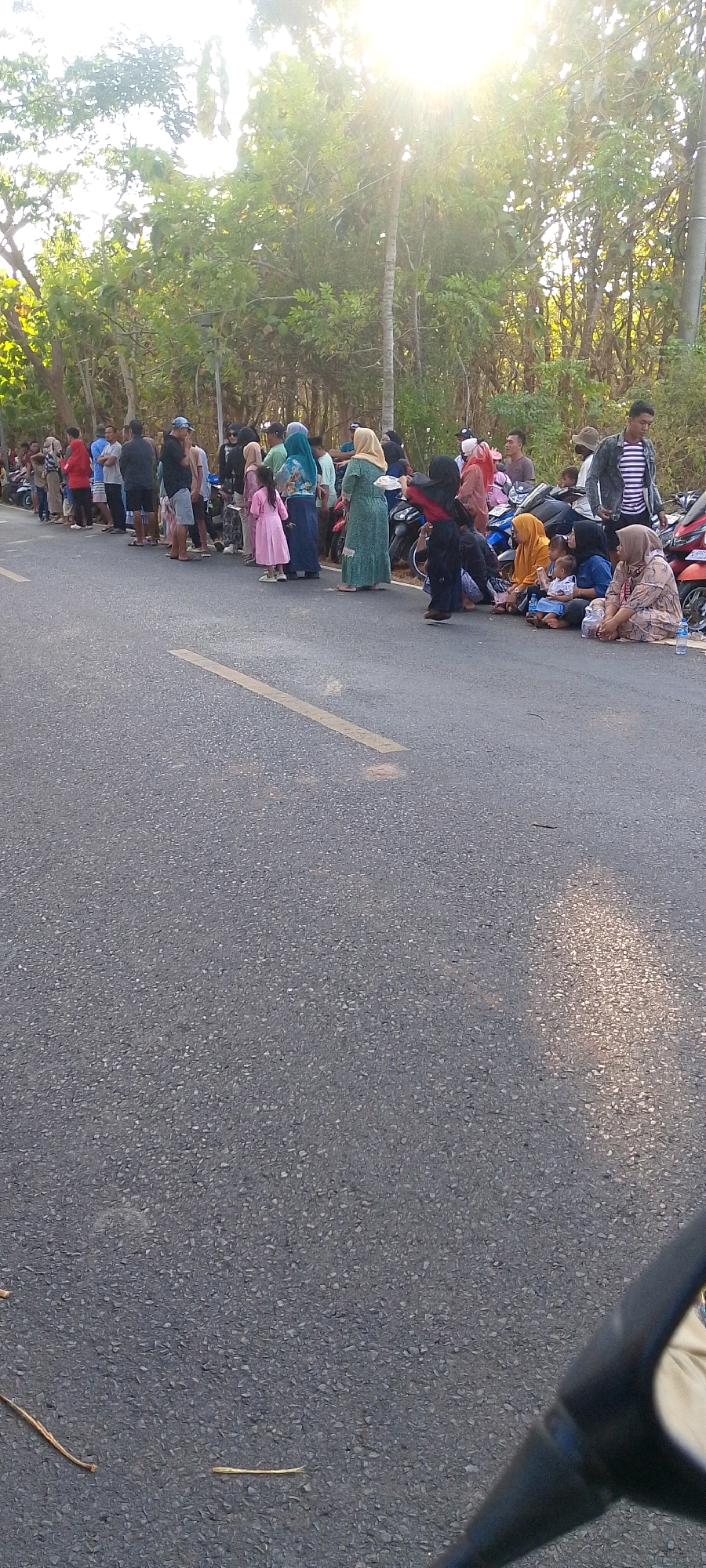 A group of people are gathered along the side of a paved road in Dempo Barat, Indonesia. The sun is shining brightly, casting a strong glare through the trees lining the road. Many individuals are dressed in casual clothing, with several women wearing headscarves. Some people are standing in a line, while others are sitting on the ground or leaning against parked motorcycles. The atmosphere appears to be one of waiting or congregating. The road itself has a yellow painted line running down the center. The background consists of dense foliage and trees.