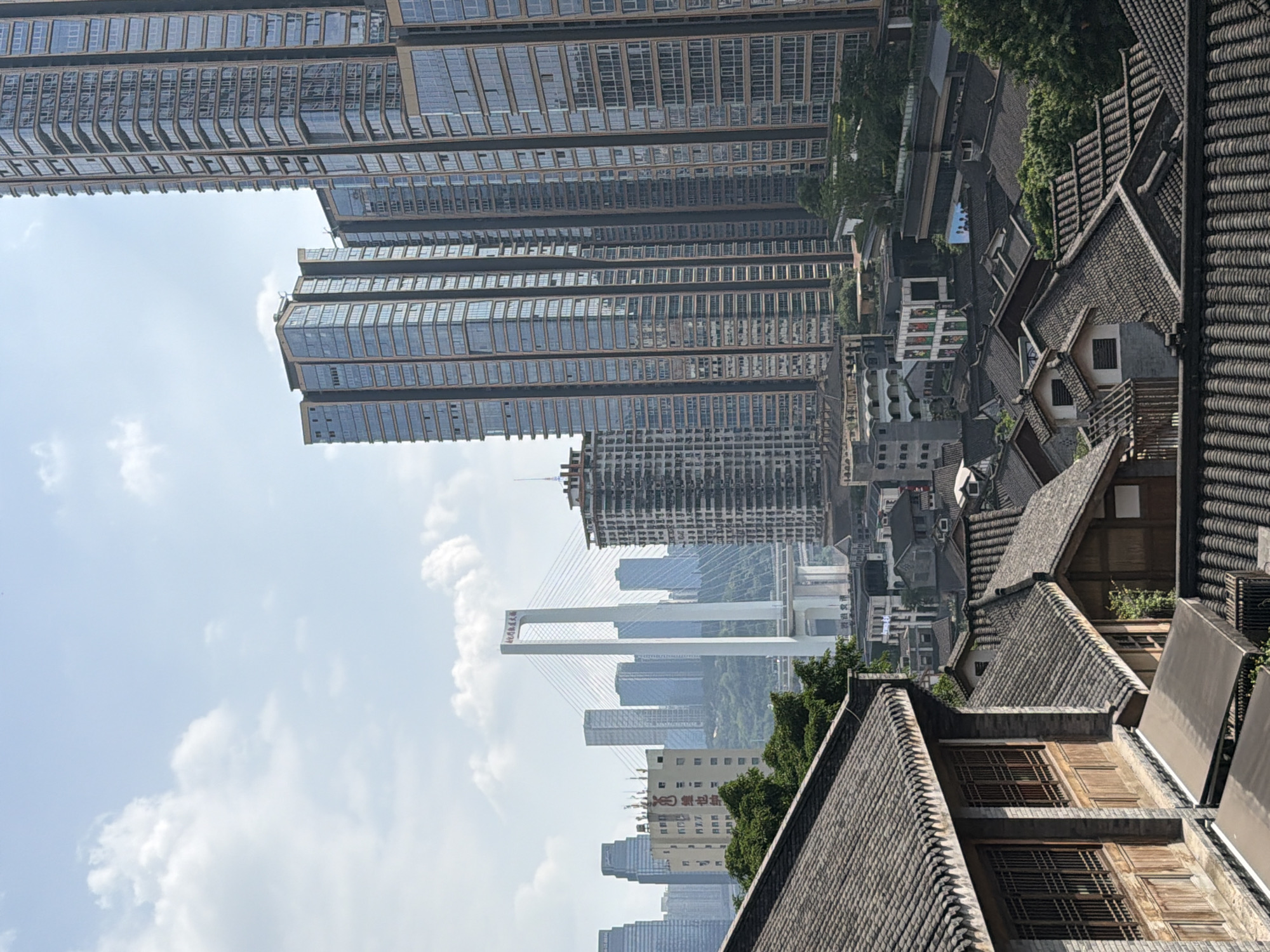 The image captures a cityscape in the Yuzhong District, China, characterized by a stark contrast between traditional architecture and modern skyscrapers. In the foreground, the tiled roofs and wooden structures of older buildings are prominent. Beyond these, a modern city unfolds, dominated by numerous tall glass-fronted skyscrapers. A large suspension bridge with white pylons spans across a body of water in the middle ground, connecting different parts of the city. The sky is partially cloudy, suggesting daytime. There are no visible people or animals in the frame. The scene depicts an urban environment with a blend of historical and contemporary elements.