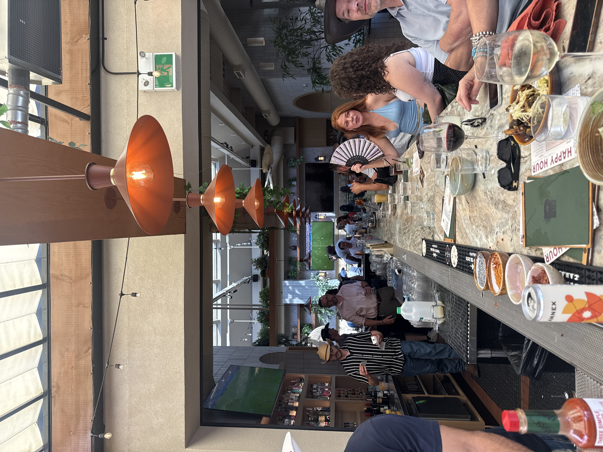 The image depicts an outdoor patio bar area, possibly in Calgary, Canada, given the presence of various elements suggestive of a bustling urban environment. The scene is focused on a long, marble-topped bar where several patrons are seated.
In the foreground, to the left, a person's arm and shoulder are visible, wearing a dark blue t-shirt. A bottle of hot sauce with a red cap is also prominently displayed on the bar. Further down the bar, a can with "ANNEX" written on it is visible, along with several small bowls containing what appear to be food ingredients or garnishes. A wine glass filled with red wine sits near the center of the image, alongside another glass containing white wine. Several other glasses, a smartphone, and sunglasses are also on the bar.
Behind the bar, a bartender wearing a striped shirt and a straw hat is interacting with a patron. Another man in a pink shirt is also standing behind the bar, holding a glass. In the background, there are shelves stocked with bottles, a television screen displaying a soccer match, and ample greenery suggesting an attempt to create an inviting atmosphere. The ceiling is a retractable awning supported by wooden beams and exposed structural elements, with industrial-style light fixtures hanging down. Several patrons are seated at the bar, engaged in conversation or enjoying their drinks. The overall impression is that of a lively social gathering at a bar and restaurant.