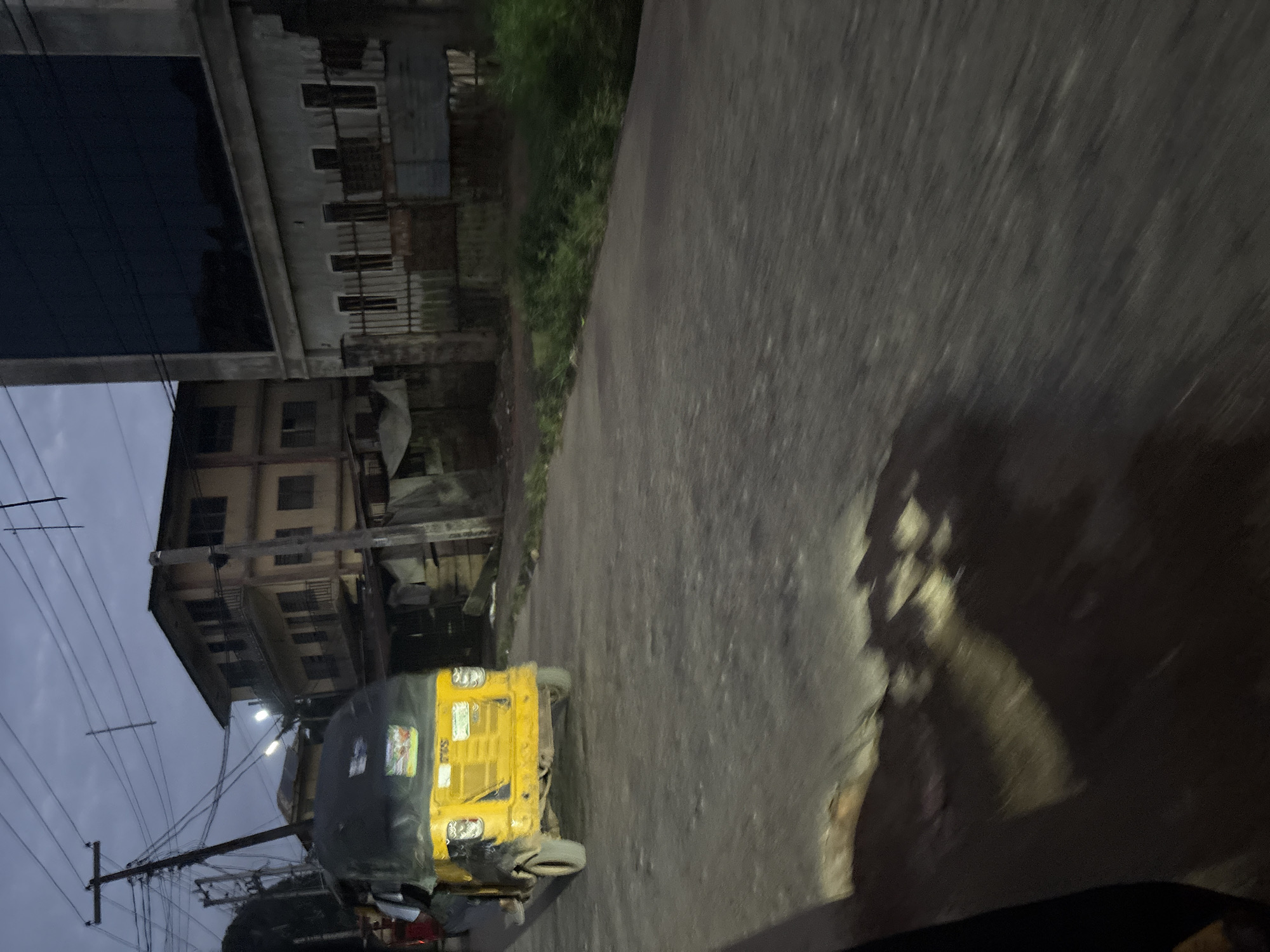 A yellow auto-rickshaw is driving on a paved road in Onitsha, Nigeria, at dusk. The auto-rickshaw is seen from the rear, with its black canopy covering most of the passenger area. The vehicle has three wheels and appears to be in motion. To the left of the auto-rickshaw, a person's legs and feet are visible, suggesting they are either driving or a passenger. In the background, there are buildings. One building is a multi-story structure with balconies and multiple windows, painted in a light brown or yellow hue. Next to it is a modern building with a dark, reflective glass facade. The sky is overcast and dark, indicating evening or twilight. Utility poles and a network of electrical wires are visible above the road and buildings. The road surface shows signs of wear, with a prominent pothole or patch in the foreground, reflecting light from an unseen source. Lush green vegetation is present along the side of the road. The scene conveys a sense of everyday urban life in a Nigerian city during the transition from day to night.