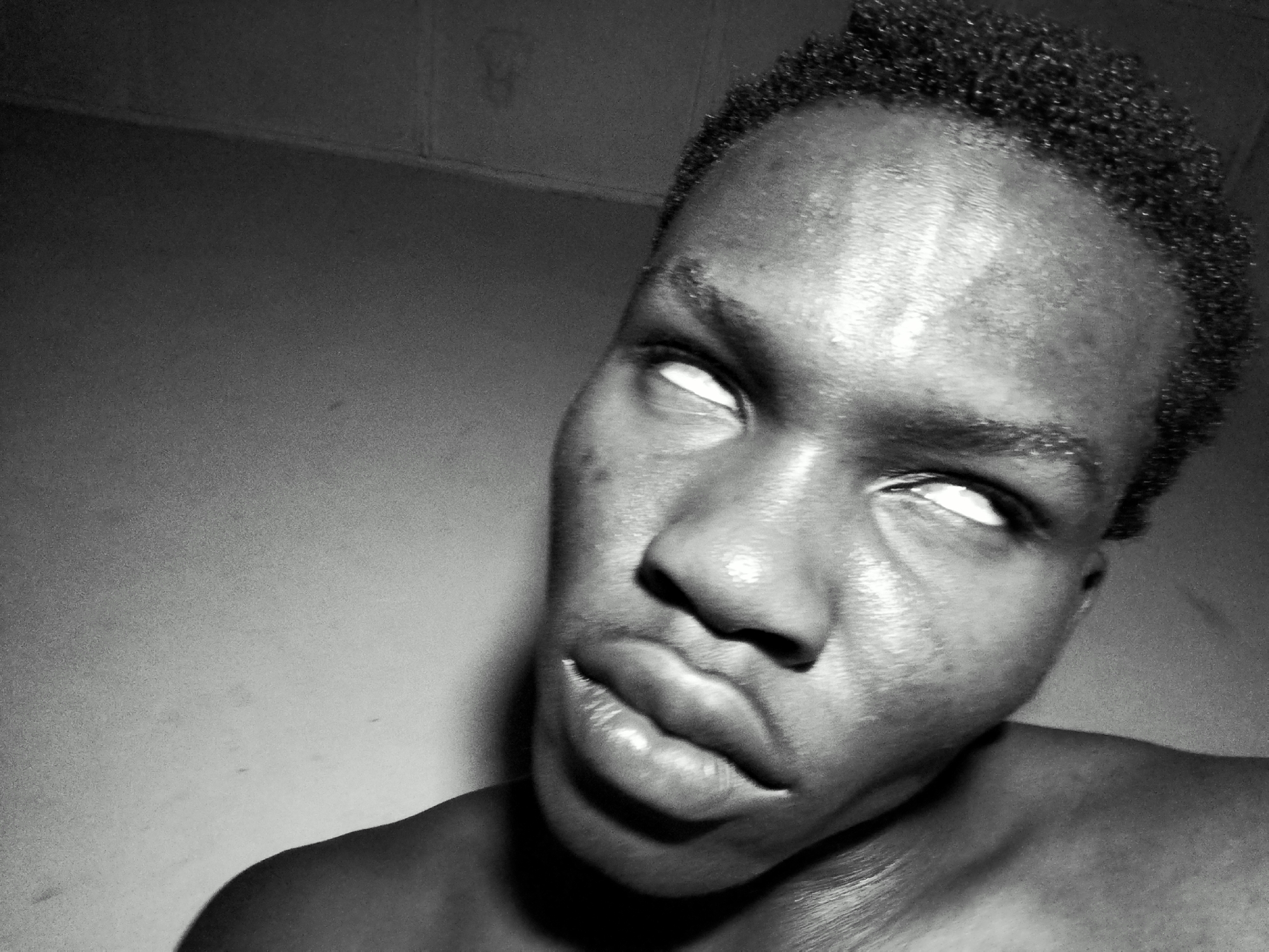 A black and white, close-up, head-and-shoulders shot shows a young Black person from the neck up, looking upwards and to the right with their eyes rolled back. The subject's skin is glistening, possibly from sweat or oil, and the lighting casts strong highlights on their forehead, nose, and cheeks. The hair is short and textured. The background is a plain, dimly lit wall. The image is framed to focus intensely on the subject's face and expression.