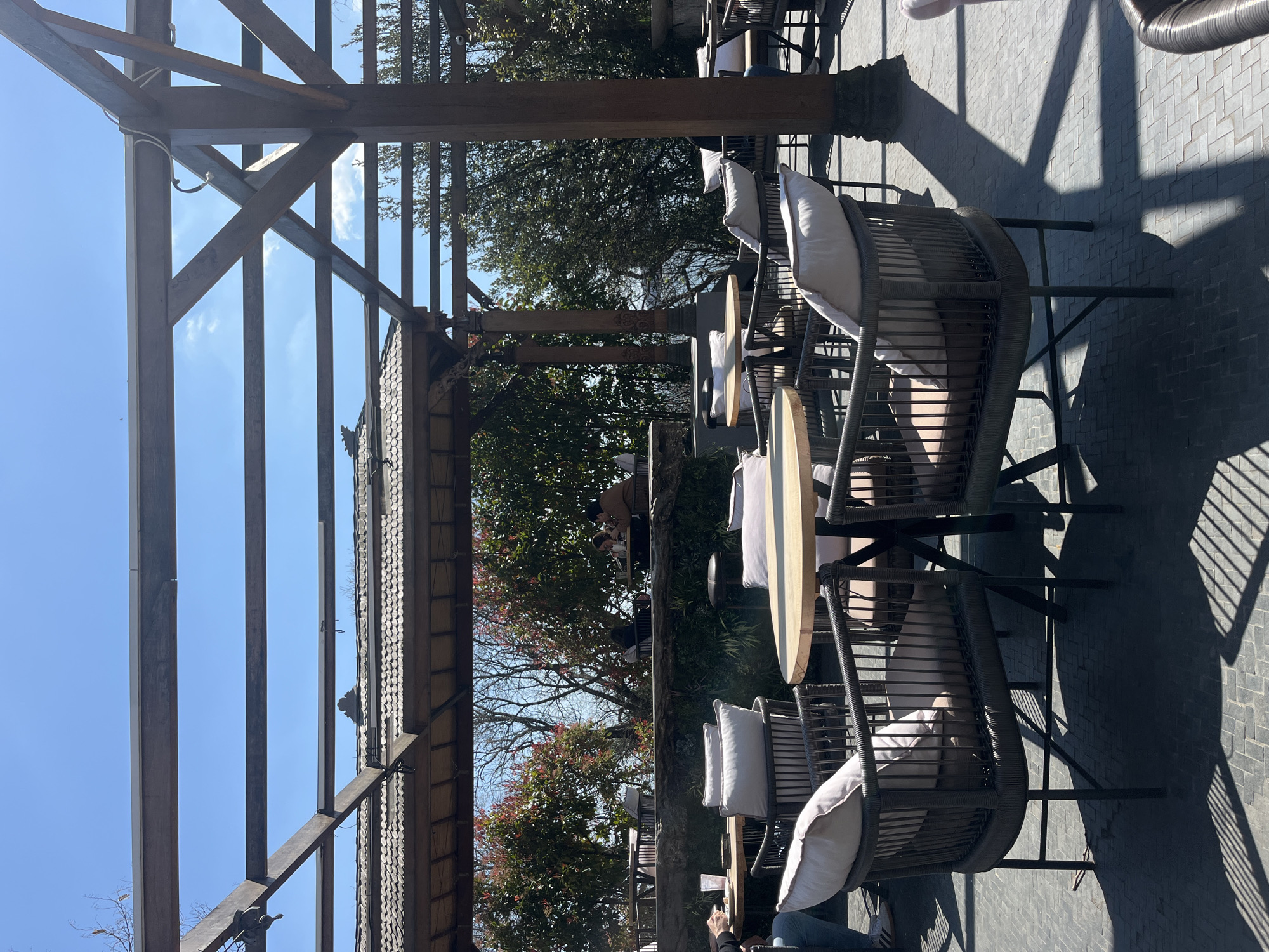 The image shows an outdoor seating area, likely in Varna, Bulgaria, featuring wicker chairs with cushions arranged around round tables. The area is shaded by a wooden pergola, which casts shadows on the paved ground. The sky is visible through the pergola's structure, suggesting a sunny day. Trees with green and red leaves can be seen in the background. People are present in the distance and a person is visible at the lower front of the image.