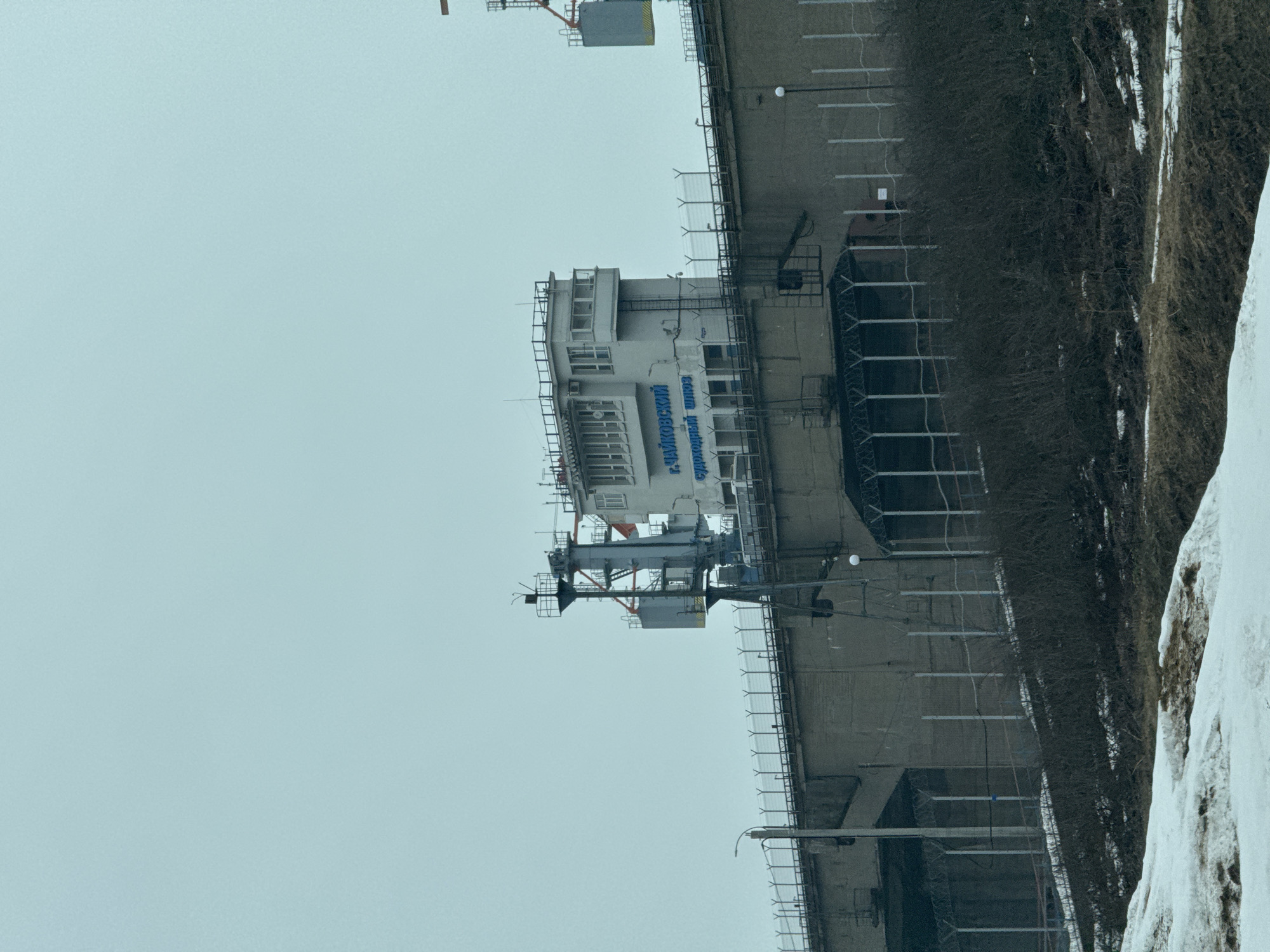 The image depicts a building in the city of Chaikovsky, Russia, specifically the Chaikovsky Shipyard. The building is a multi-story structure with a control tower-like section. The building is gray. There is a parking lot in front of the building. The sky is overcast. There is some snow on the ground.