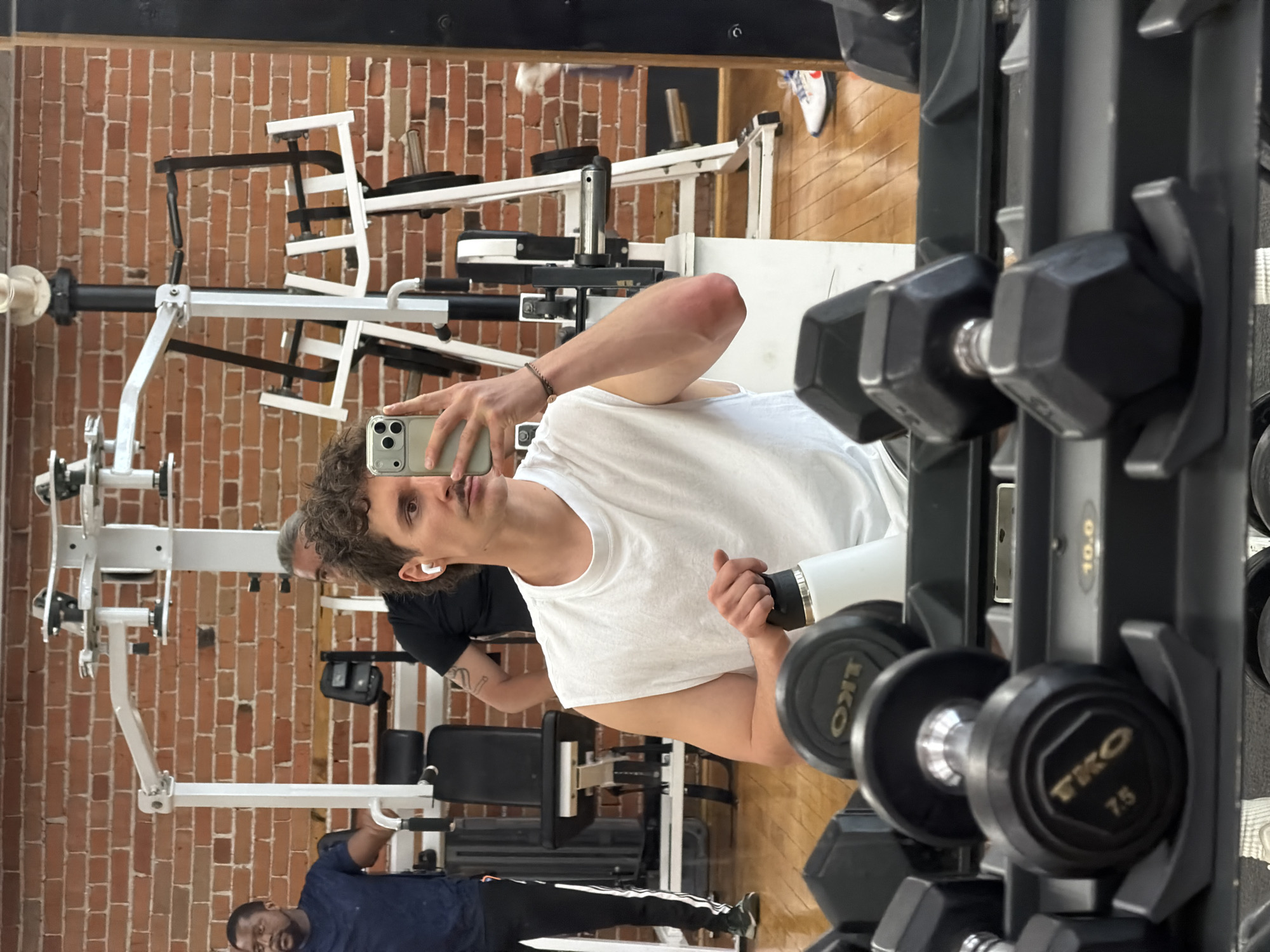 The image shows a man taking a selfie in a gym. He is wearing a white t-shirt and holding a white water bottle. He has curly hair and is wearing wireless earbuds. In the background, there are other people working out on weight machines, and there are racks of dumbbells. The gym has a brick wall. The environment appears to be a commercial gym. The details do not provide enough information to determine if it is in Montreal, Canada.