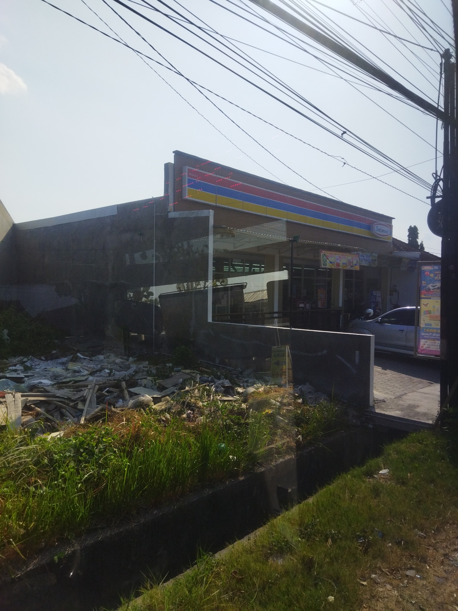 The image shows a daytime exterior view of a convenience store, possibly an Indomaret, in the city of Begajah, Indonesia. The store features a prominent red, blue, and yellow striped awning. To the left of the store, there is a large pile of construction debris and discarded materials. Tall green grass and a dark drainage ditch are visible in the foreground. A silver car is parked in front of the store, and a person on a motorbike is also present. Overhead, a dense network of electrical wires crisscrosses the sky. The scene suggests a somewhat unkempt environment surrounding a commercial establishment.
