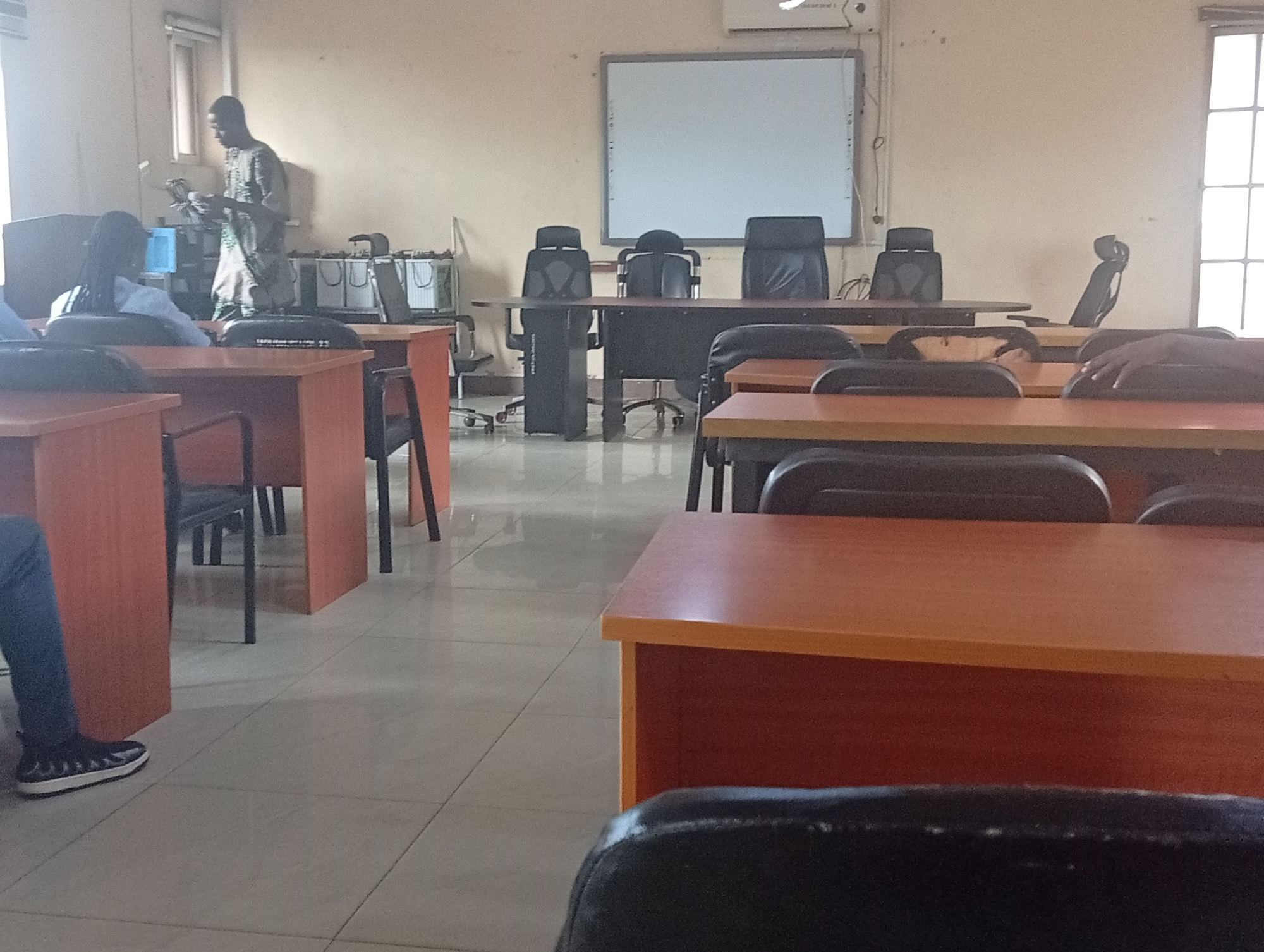 The image depicts an indoor scene, possibly a classroom or conference room in Ijanikin, Nigeria. Desks are arranged in rows, with black chairs positioned at each desk. A man stands at the front of the room, holding papers and looking down, appearing to read. Other people are seated at the desks. In the background, there's a table with three black office chairs, and a whiteboard is mounted on the wall. A window is visible on the right side of the frame. The room appears to be lit by overhead lights and natural light from the window.