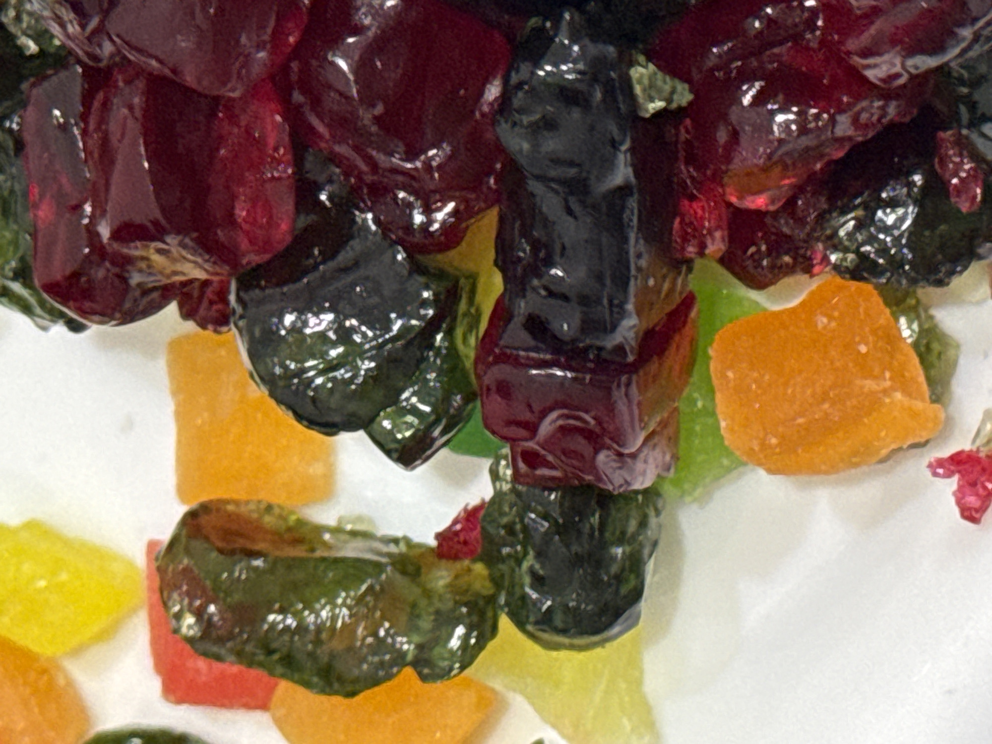 A close-up shot depicts a cluster of colorful candied fruit pieces. The dominant colors are deep red, dark green, orange, and yellow. Some pieces are glossy and appear to be covered in a sugary coating, while others are matte. The shapes of the fruit pieces are irregular. The background is plain white. The location is not discernible from the image. There are no people present.
