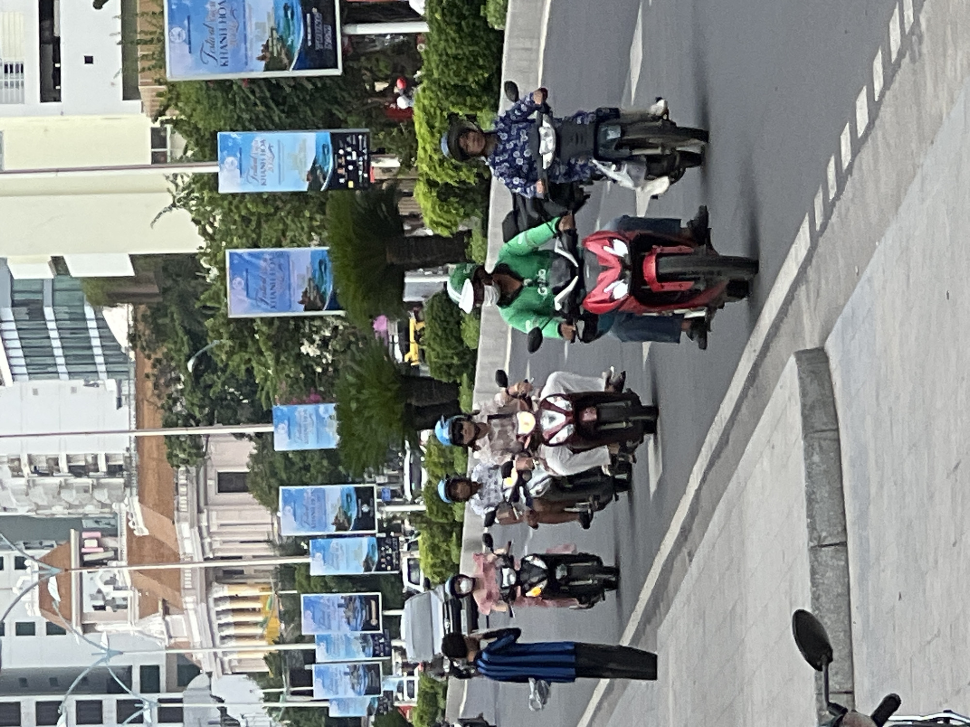 The image depicts a street scene in Nha Trang, Vietnam, likely along the coastal road given the presence of palm trees and buildings that suggest a resort city. In the foreground, several people are riding motorbikes. A person in a blue and black striped shirt stands on the sidewalk looking at their phone. Further down the street, a woman in a pink dress rides a black motorbike, followed by two individuals on another motorbike, one wearing a plaid shirt and the other a checkered shirt. On the right side of the road, a person wearing a green Grab uniform and a face mask rides a red motorbike, with another person on a motorbike behind them wearing a blue floral patterned shirt. In the background, various buildings, including a light-colored building with columns and a brown-tiled roof, are visible. Several tall lampposts with banners advertising "Festival Khanh Hoa 2023" are also present. The road is paved and shows lane markings. Traffic appears moderate, with cars and other motorbikes visible in the distance. Lush green trees and shrubs line the street.
