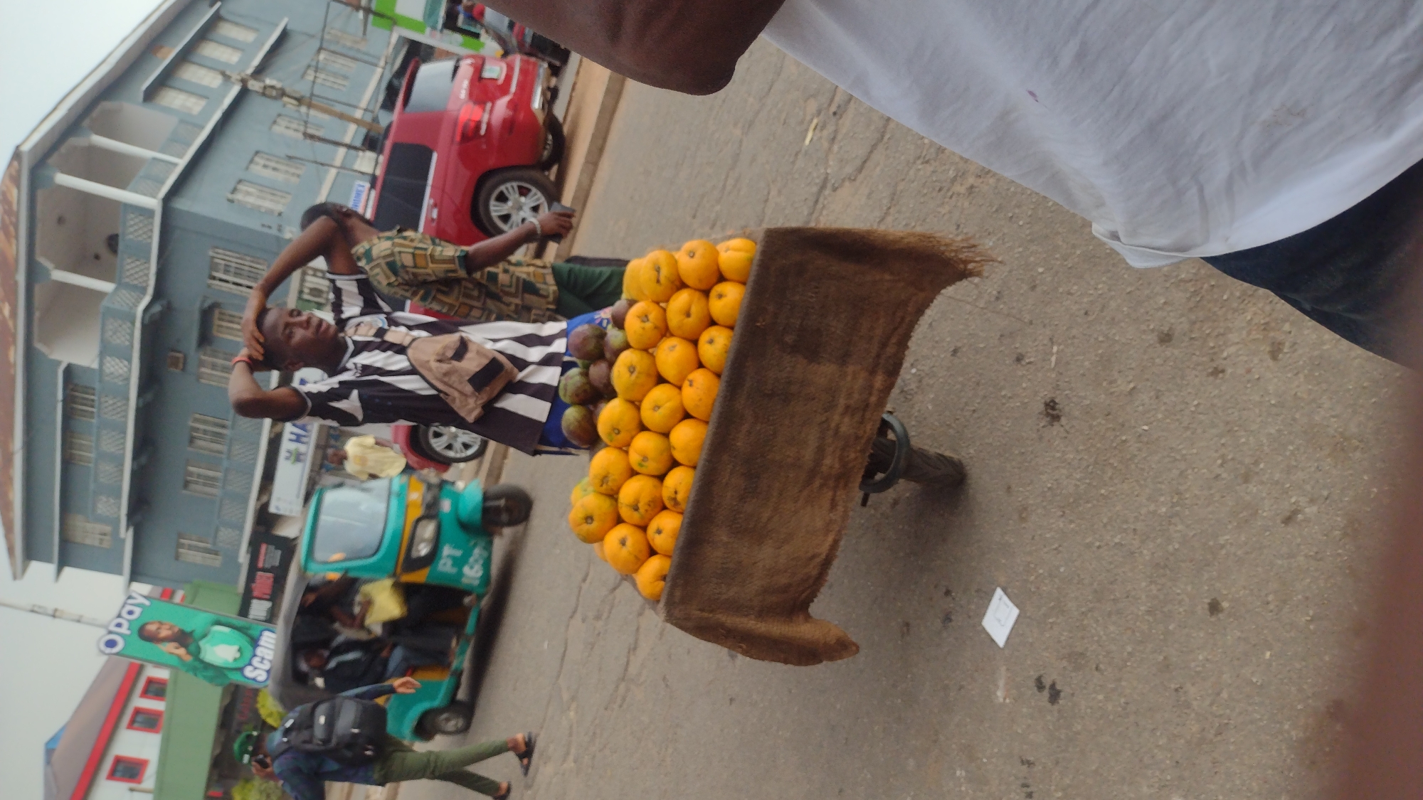 A vendor in Jos, Nigeria is displaying his produce on a cart. The produce consists of oranges and mangoes. The vendor is wearing a black and white striped shirt with a brown cross-body bag. He is holding his head with both hands. In the background, there is a building with a blue facade and a red SUV. An advertisement for Opay is on a passing green tricycle. A person in khaki pants walks toward the tricycle. A piece of white paper lays on the street. An unidentified person is partially shown in the top right.