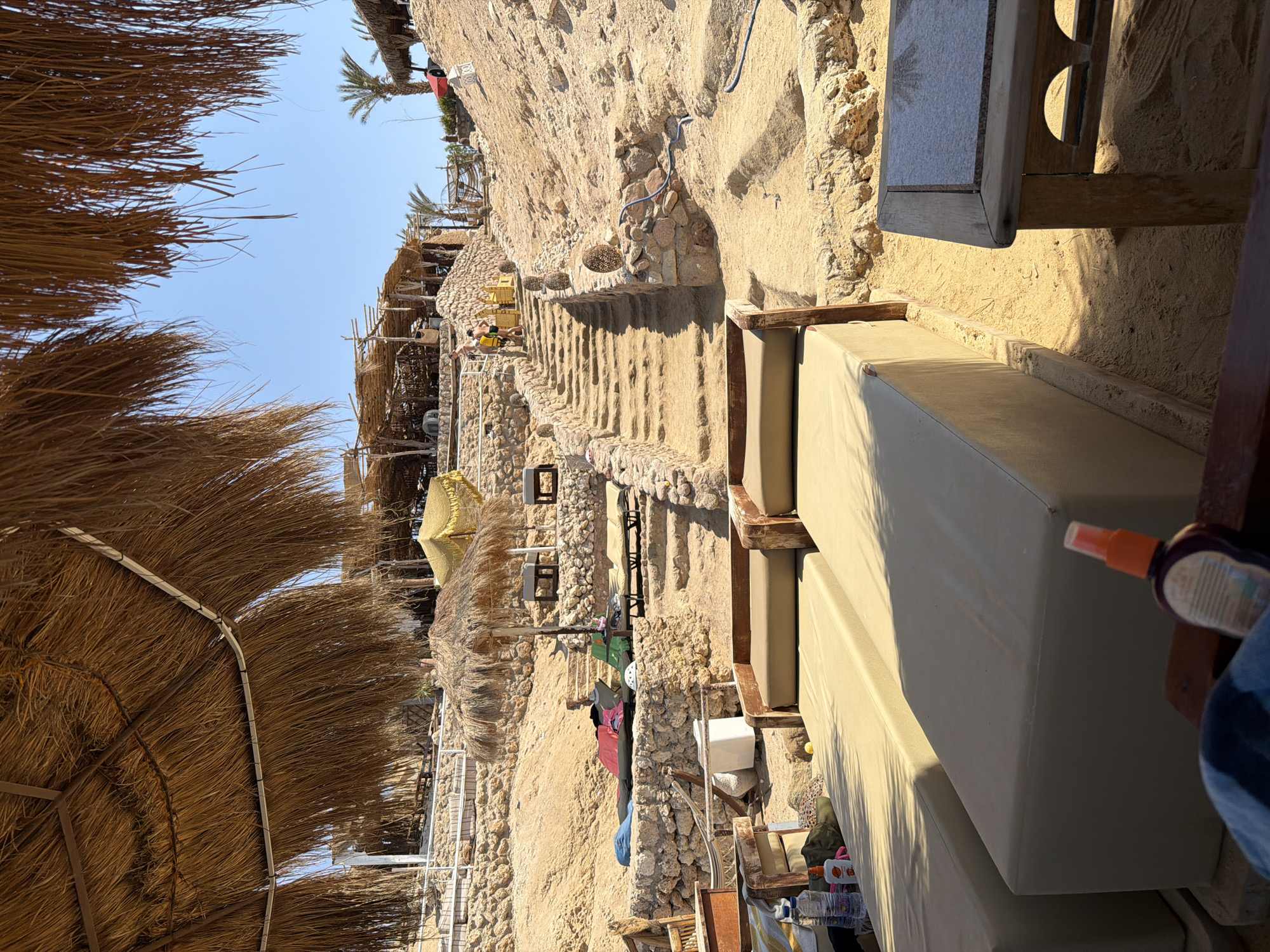 The image depicts a beach resort scene in Sharm El Sheikh, Egypt, characterized by a sandy incline and a clear blue sky. In the foreground, two sun loungers covered in light beige upholstery are prominently placed on the sand, partially obscured by a wooden frame. To the right, a small wooden table with a granite top sits adjacent to the loungers. The scene further back reveals a stone staircase ascending a sandy hill, with more sun loungers and thatched umbrellas scattered along the path. Several structures with thatched roofs are visible further up the incline, suggesting a restaurant or bar area. Two individuals are seen walking up the stairs. The environment is designed for relaxation and leisure, with a focus on outdoor seating and sunbathing amenities.