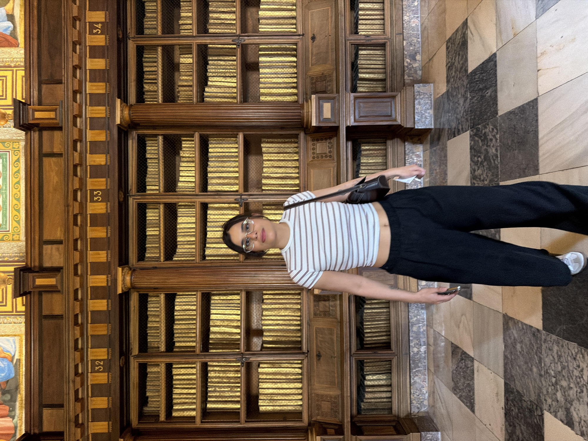 A young woman with dark hair and glasses stands in the middle of a room in the library of San Lorenzo de El Escorial, Spain. She is wearing a white and brown horizontally striped crop top and black wide-leg pants. She is carrying a dark brown purse over her left shoulder and holding a white object in her right hand. Behind her, floor-to-ceiling bookshelves filled with gold-spined books line the walls. The shelves are ornately carved wood, with decorative elements and numbered sections. The floor is a patterned marble in shades of beige, gray, and black. The room is well-lit, with overhead lighting highlighting the books and the woman. The overall impression is one of grandeur and historical significance.