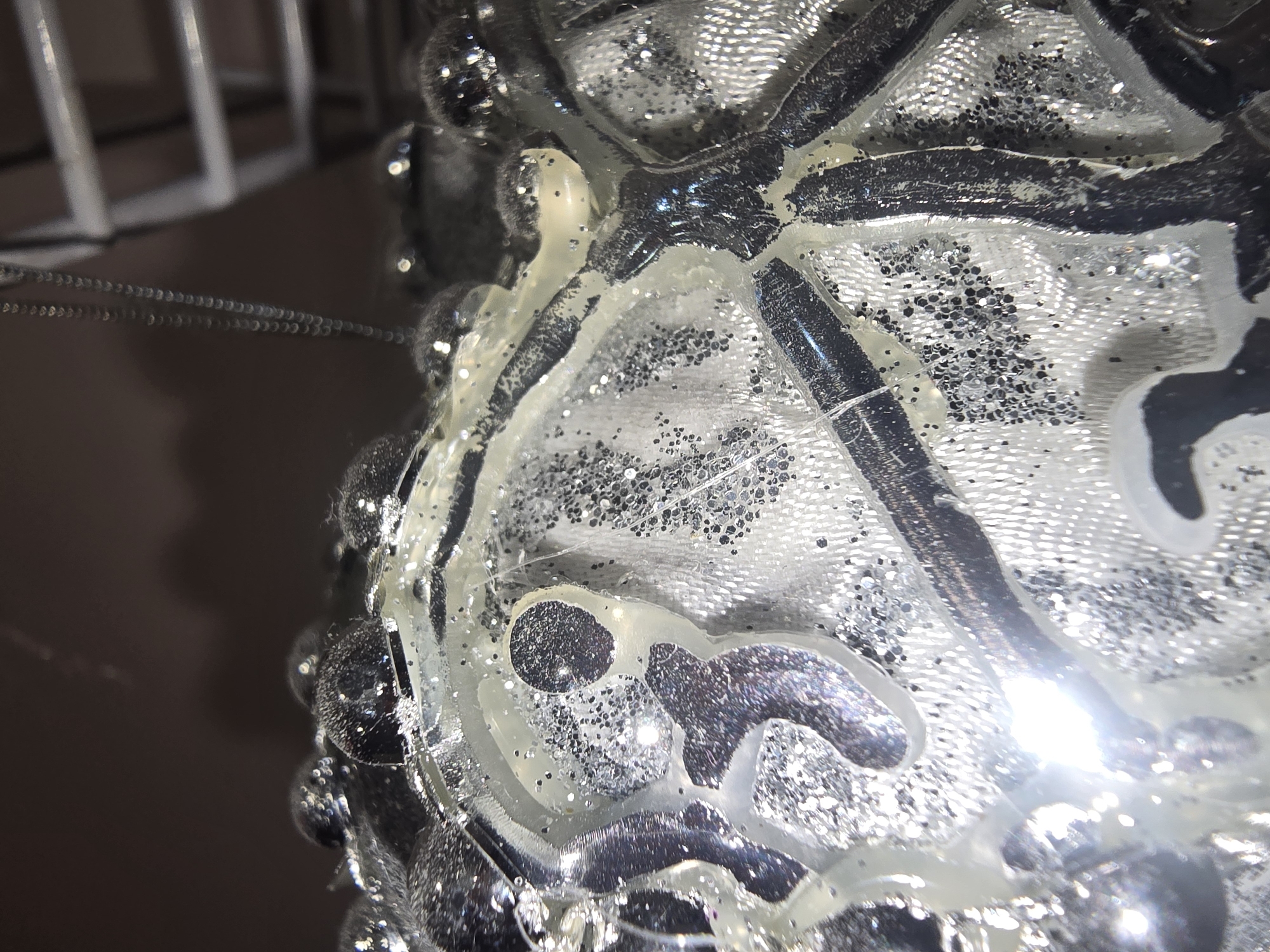 The image displays a close-up, macro view of a decorative object, likely a Christmas ornament. The object is primarily silver and clear, with a textured, slightly translucent surface covered in glitter. Darker, raised elements, possibly depicting a floral or abstract pattern, are visible on the surface, embedded within a clear, gel-like substance that also contains glitter. Some spherical, raised bumps, also adorned with glitter, are present along the edge of the object. The lighting creates strong reflections and highlights, particularly on the glossy surfaces and the glitter. In the background, a blurry chain-like object and what appear to be shelves or a ladder are dimly visible, suggesting an indoor environment. The image does not provide any information that would allow for its location to be identified as Ulan-Ude, Russia, or any other specific geographical location.
