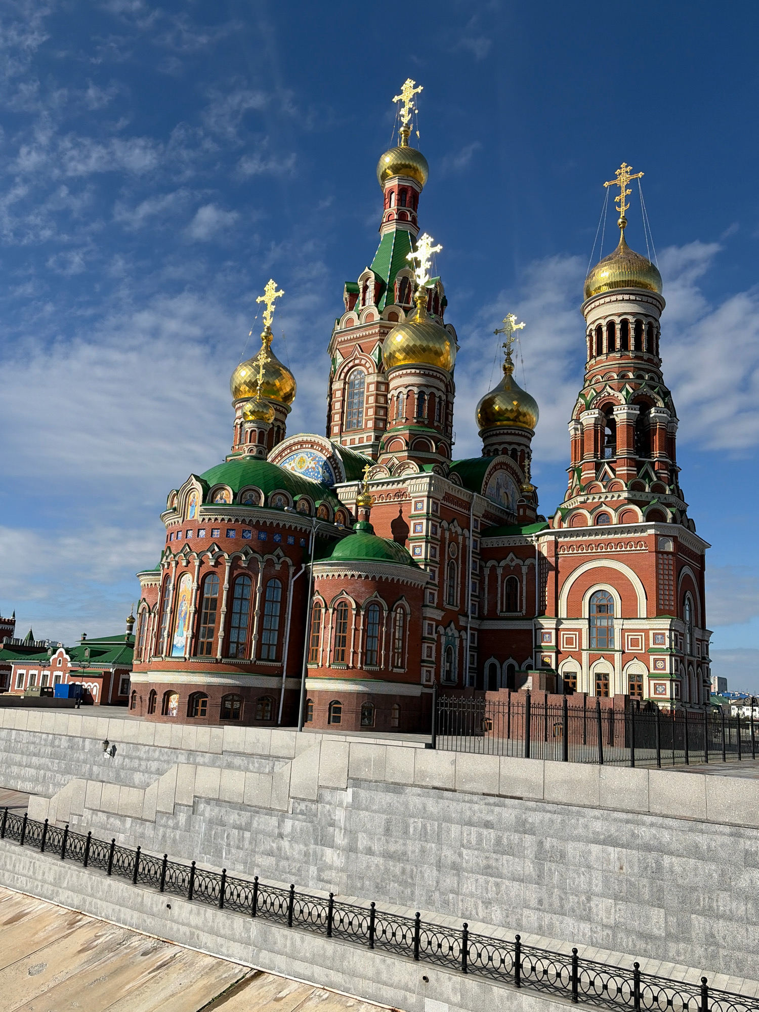 The image shows the Annunciation Cathedral, a prominent Orthodox church located in Yoshkar-Ola, Russia. The cathedral is a large, red brick building with multiple green-domed roofs and golden cupolas topped with crosses. The architecture is ornate and features intricate detailing in white stone and colorful mosaics. The cathedral is situated in a public space, with a paved walkway and a black metal railing in the foreground, suggesting it is along a waterfront or embankment. Several steps made of light-colored stone lead up to the building's base. A black metal fence separates the walkway from the cathedral. In the background, more buildings with green roofs and red brick are visible, along with trees and a clear blue sky with scattered clouds. Two figures are present near the steps, appearing to be walking or standing. The overall scene depicts a bright, sunny day in a well-maintained urban environment focused on the majestic cathedral.