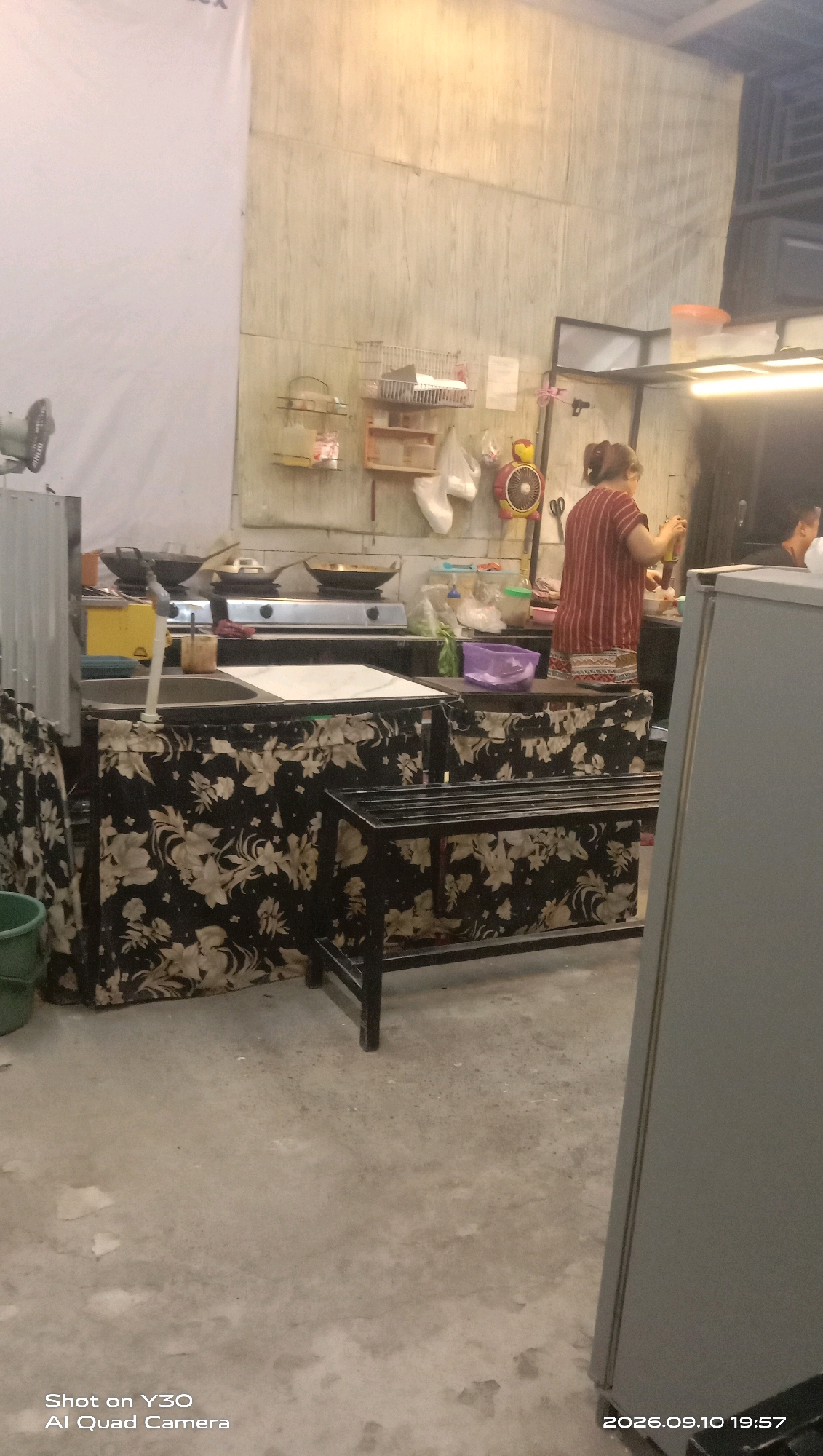 A street food stall in Karangankidul, Indonesia, is captured in a medium shot. The stall is set up in an open area, likely a sidewalk or street, with a concrete floor. The stall features a cooking area with a gas stove, pots, and pans. A woman wearing a red and white striped shirt and shorts is standing behind the counter, preparing food. A shelf unit with various containers and bags is mounted on the wall behind her. To the left of the cooking area, there is a sink and a yellow object, possibly a cooler or storage container. The front of the stall is covered with a black fabric adorned with a floral pattern. A metal rack is placed in front of the stall. In the background, a large white sheet or banner hangs on the wall. The lighting appears to be artificial, suggesting it might be evening or the stall is under a covered area.